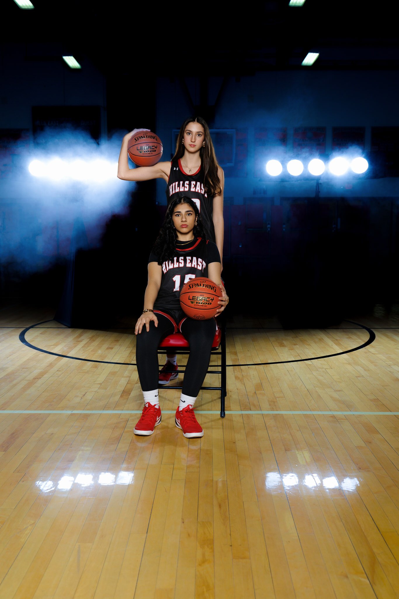 Two young women in basketball uniforms sitting in a basketball court with bright lights and smoke in the background. One standing behind the other, holding a basketball on her shoulder, and the other seated holding a basketball.