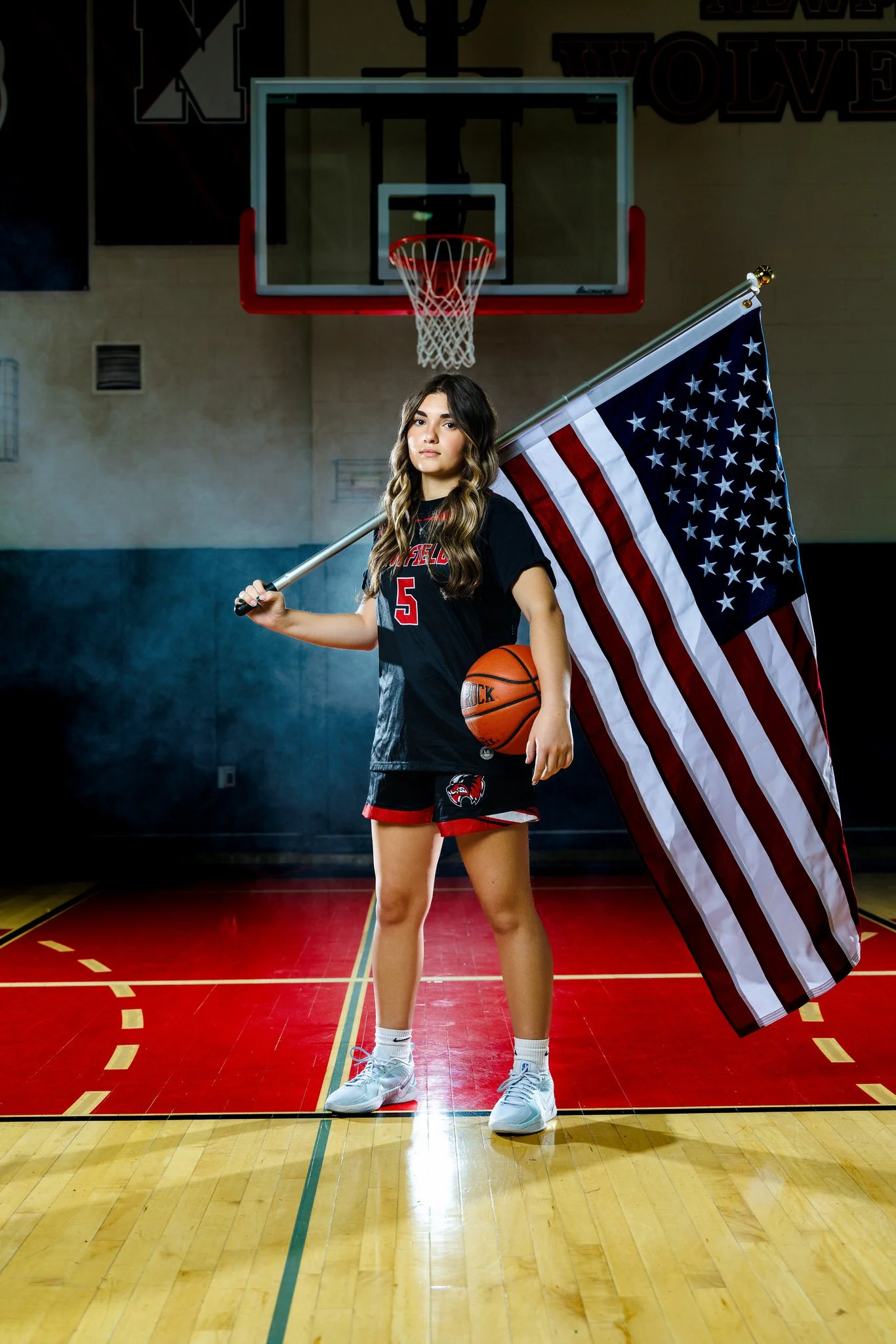 A young female basketball player standing on a basketball court, holding an American flag and a basketball. She is wearing a black and red sports uniform with the number 5, white sneakers, and has long wavy hair. The background includes a basketball 