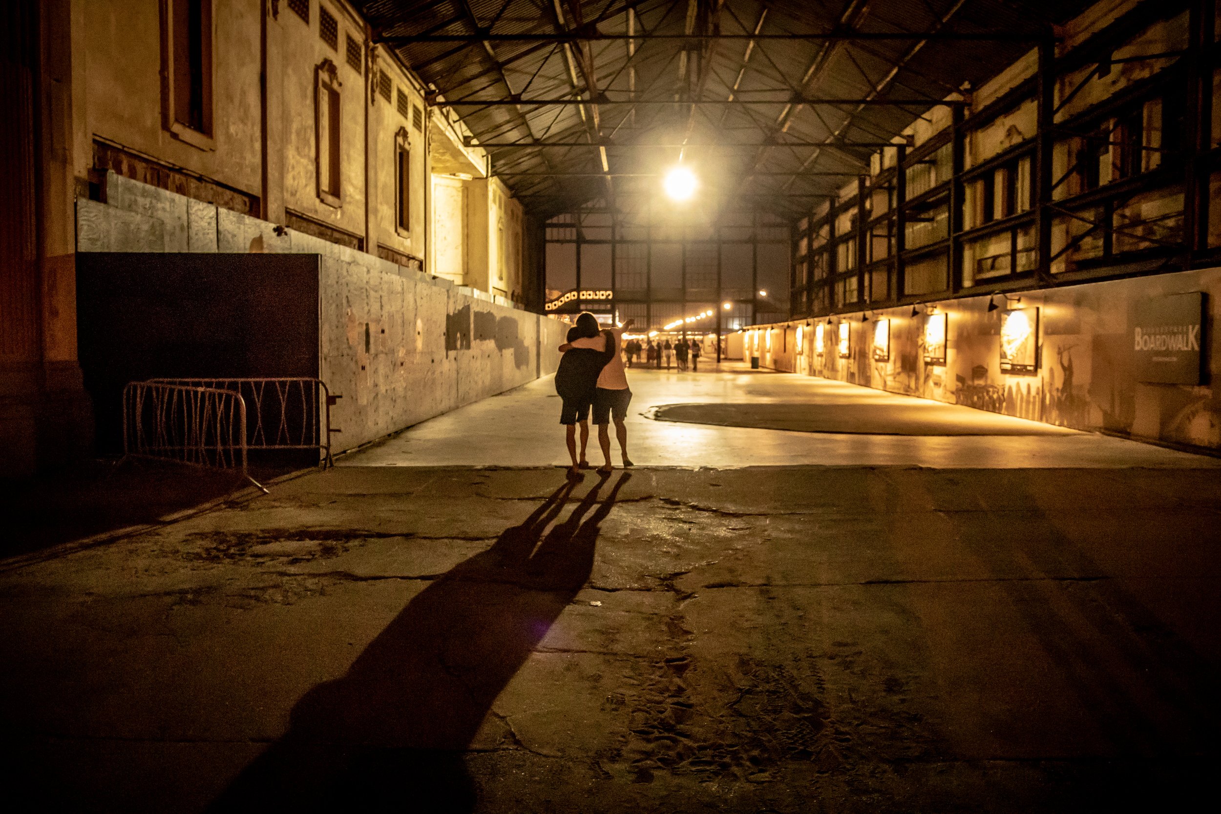 Dylan and Hayden (brothers) hugging in an industrial-style indoor space illuminated by warm yellow lighting, with a high ceiling, metal beams, and a painted wall. (Asbury Park, NJ)