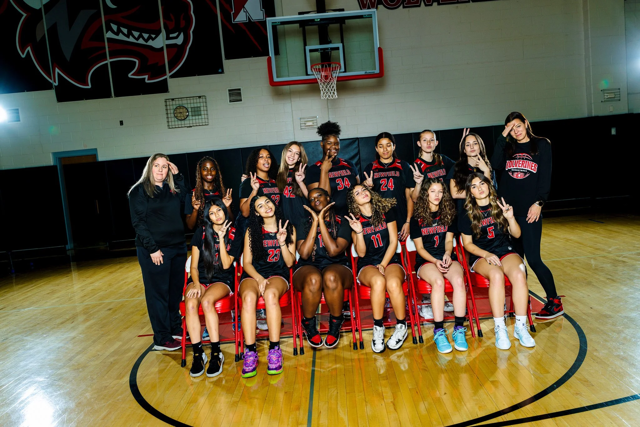 A girls' basketball team posing for a photo on a basketball court inside a gymnasium. The team is wearing black uniforms with red and white accents, and the word "Newfield" on the front. They are standing and sitting, making various hand gestures, wi