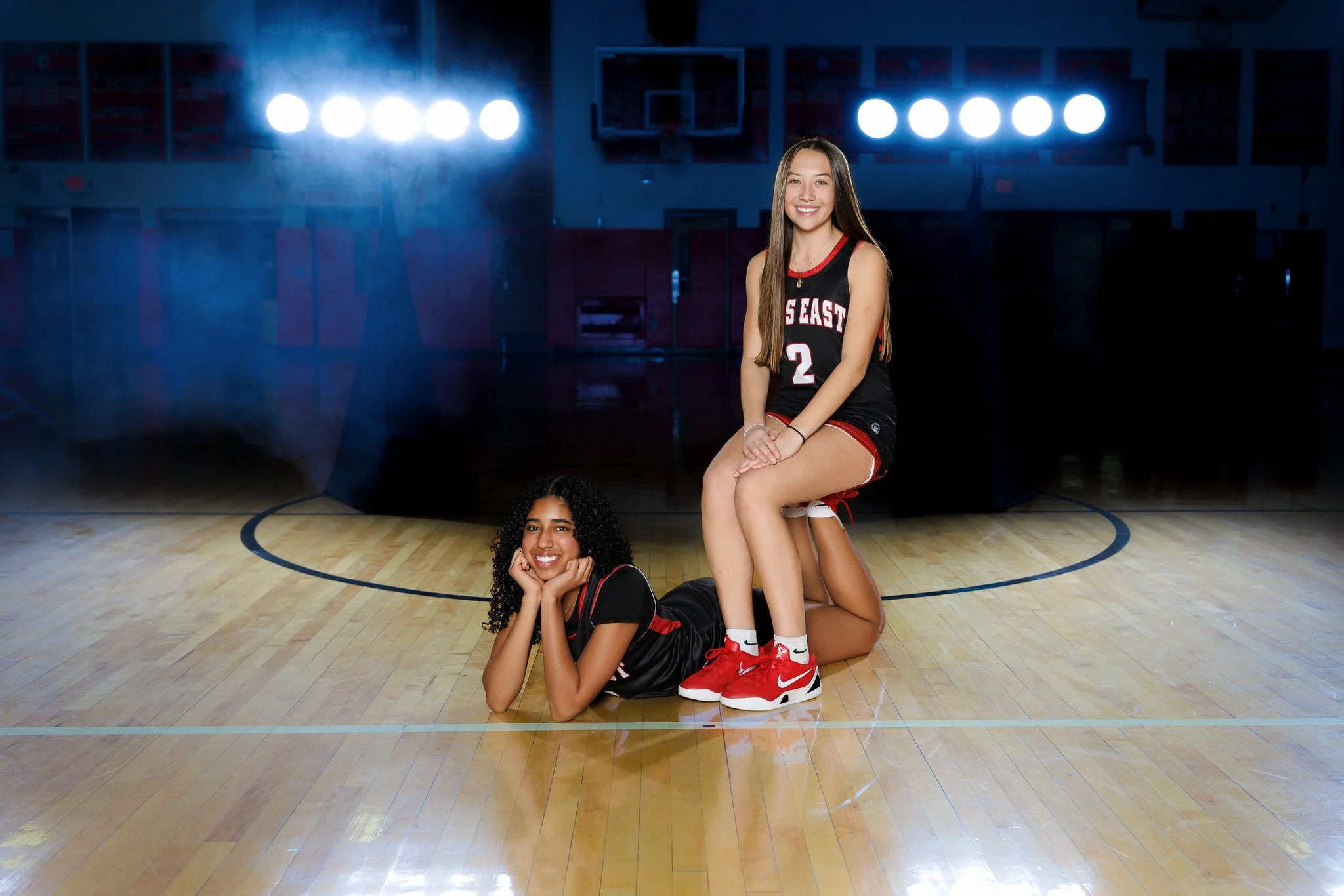 Two young female basketball players in black and red uniforms, one sitting on the floor with her head resting on her hands and the other sitting on her teammate's knees, both smiling, in a gymnasium with bright overhead lights in the background.
