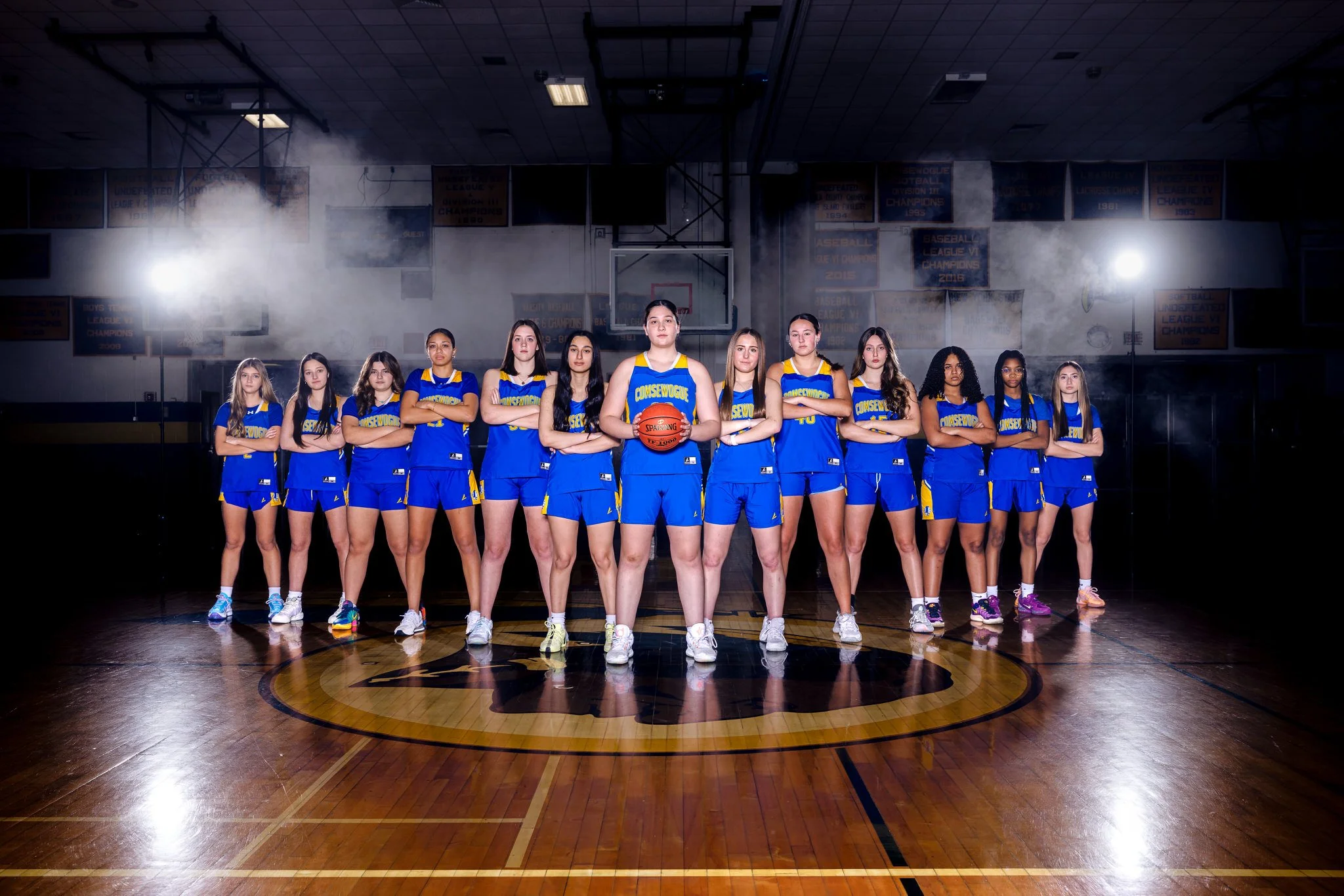 A girls' basketball team in blue and yellow uniforms standing in a sports gymnasium, with one girl holding a basketball, posing for a team photo.