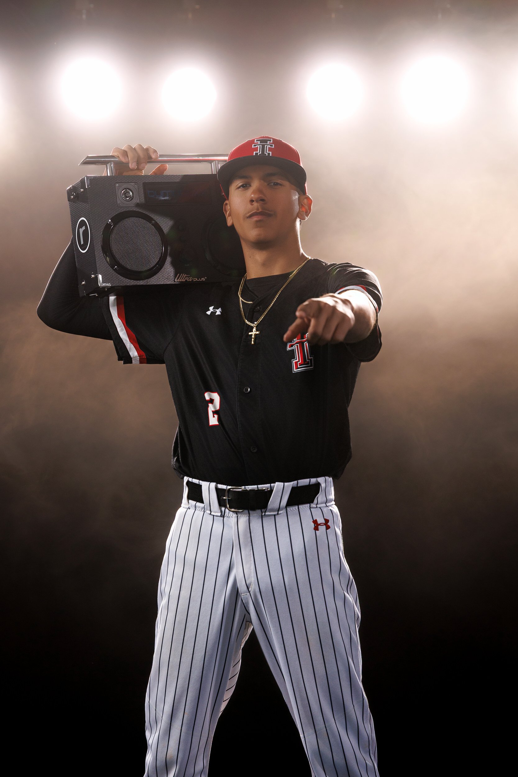 Young male baseball player is posing confidently, pointing at the camera, holding a portable speaker on his shoulder, wearing a black baseball jersey, pinstriped pants, a red cap, and a gold chain with a cross.