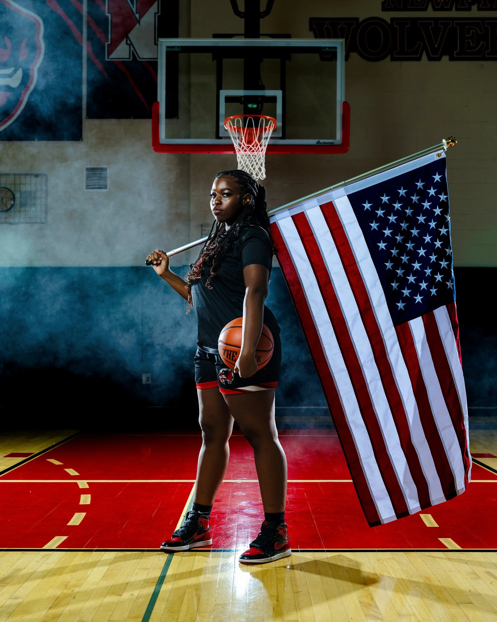 A female basketball player standing on an indoor basketball court holding an American flag over her shoulder and a basketball in her other hand, with a basketball hoop in the background.
