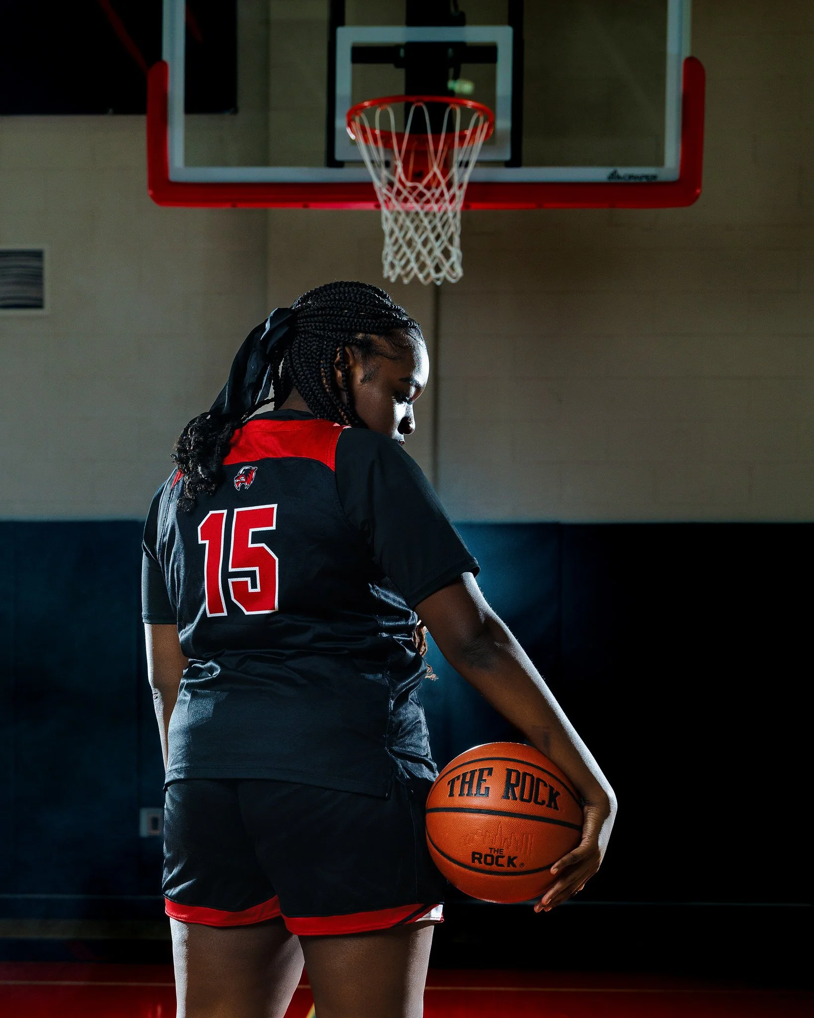 A female basketball player wearing a black and red uniform with the number 15, holding a basketball titled 'The Rock,' standing in a gymnasium with a basketball hoop overhead.