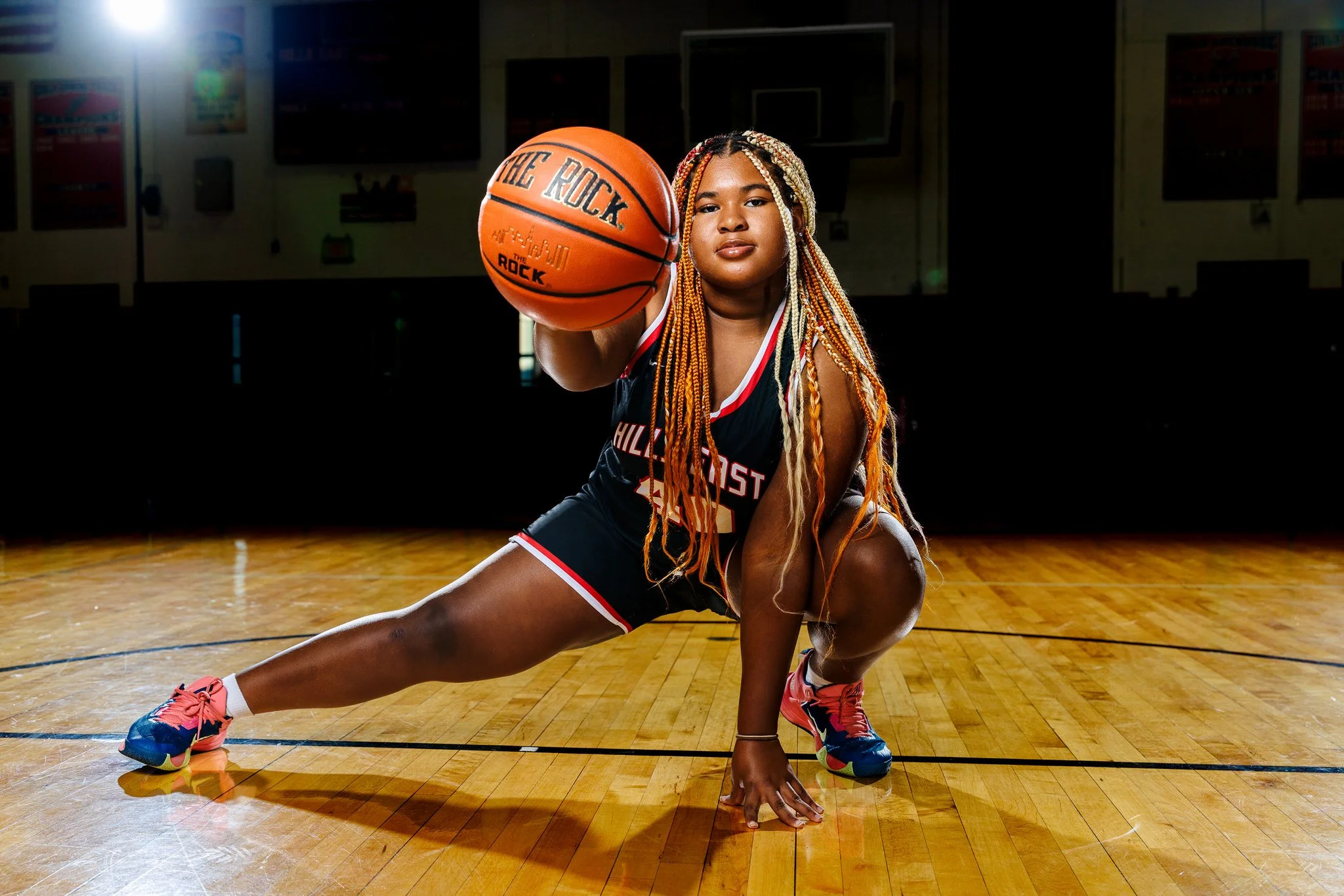 Young female basketball player in black and red uniform kneeling on a gym floor, holding a basketball towards the camera with her right hand, with her left hand and knee on the ground, in a low lunge position.