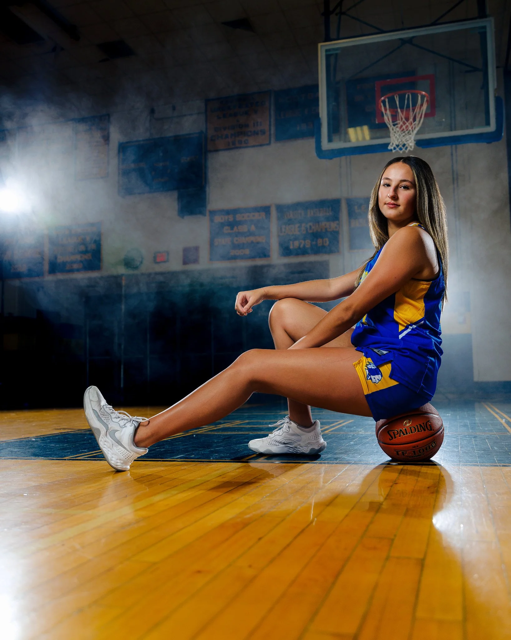 A young female basketball player sitting on the court with a basketball under her arm, in a gymnasium with banners on the wall and a basketball hoop in the background.