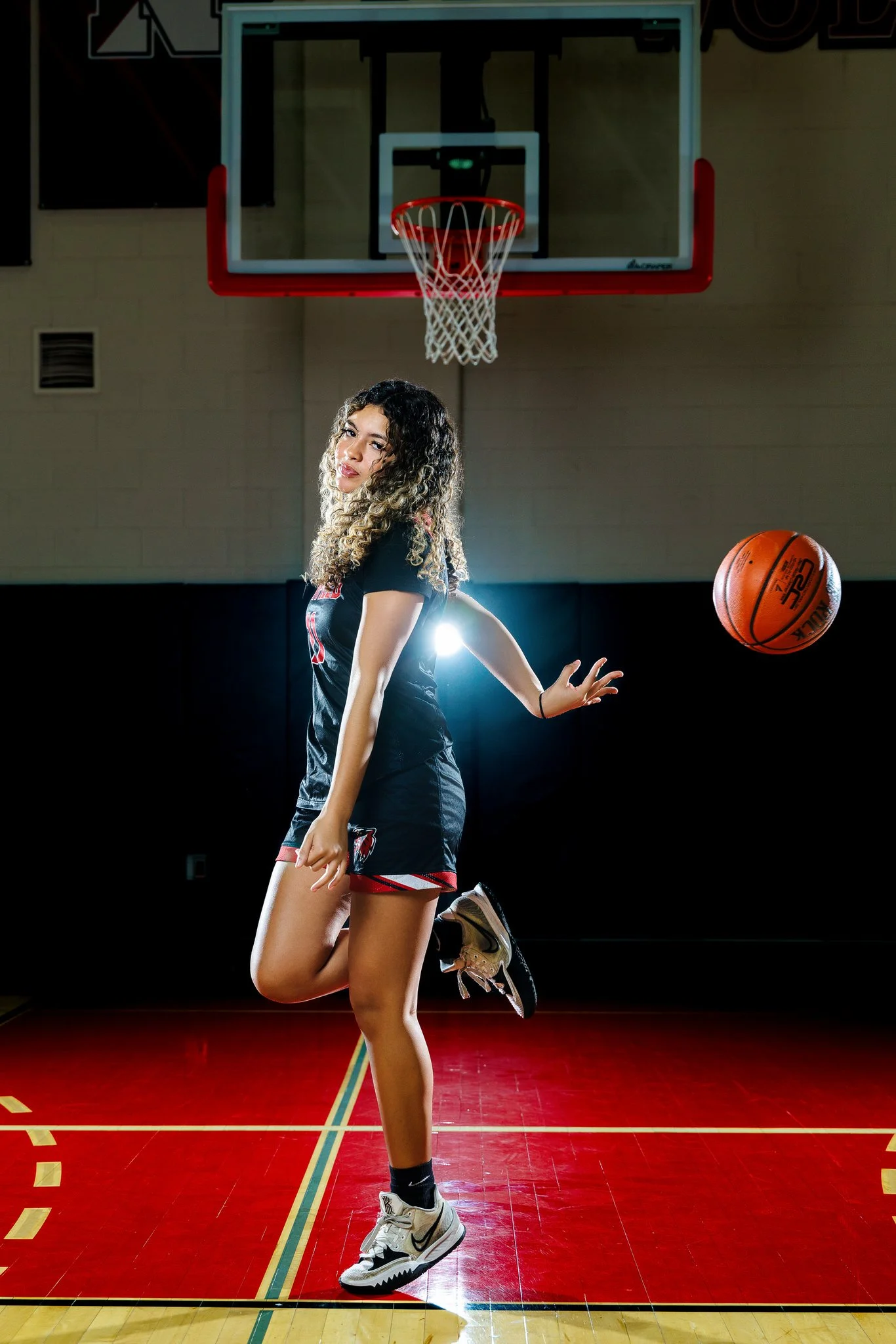 A young woman with curly hair dressed in a black and red basketball uniform is on an indoor basketball court, about to make a shot or perform a move with a basketball in mid-air near her right hand. She is standing on her left leg with her right knee