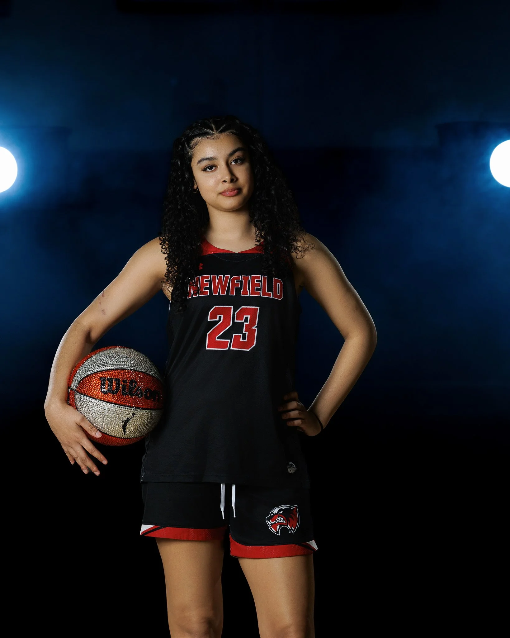 A young female basketball player with curly hair, wearing a black and red uniform with the number 23 and the team name 'Newfield,' holding a basketball, standing confidently against a dark background with two bright lights.