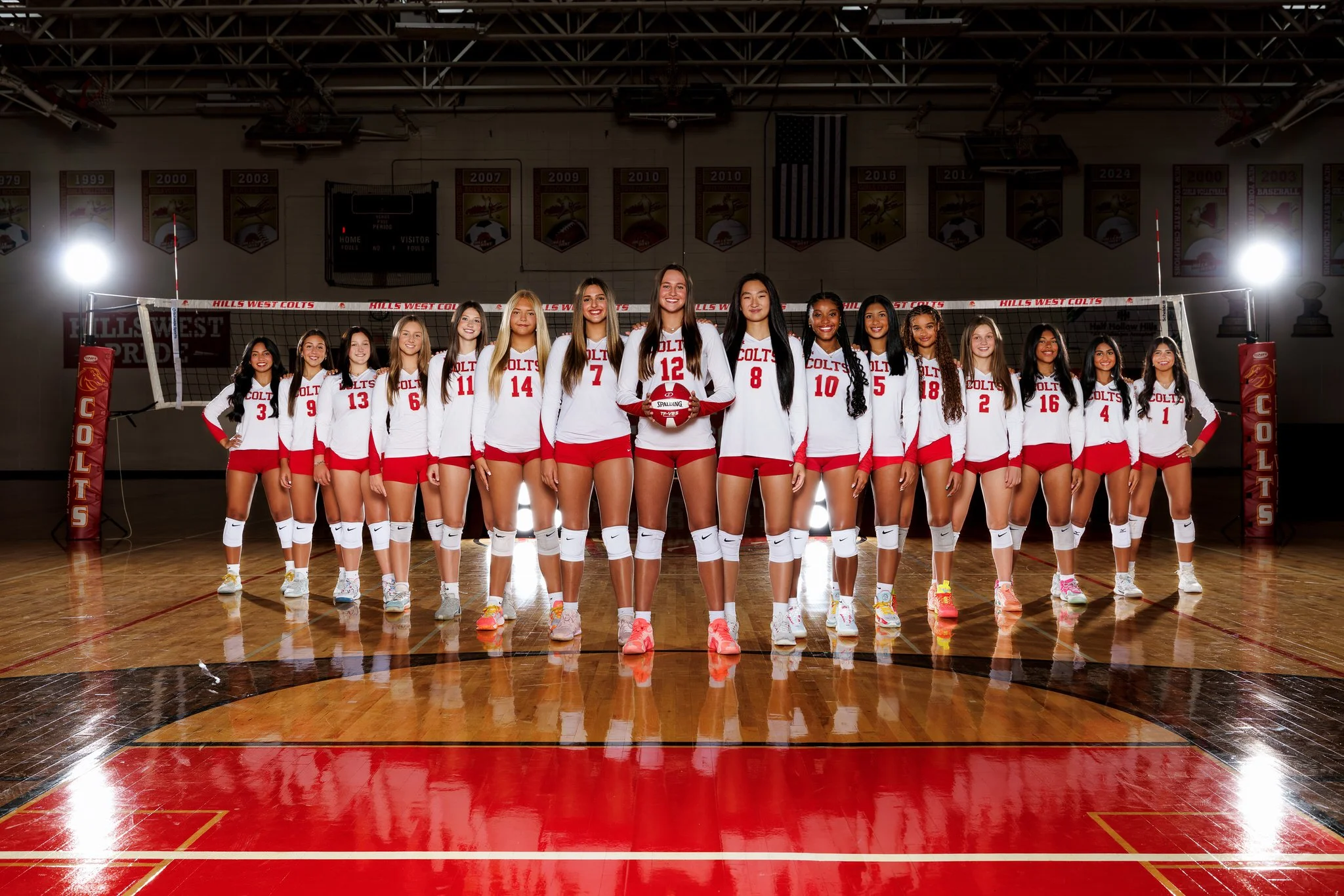 A girls' volleyball team in red and white uniforms standing together on a gymnasium court, posing for a team photo with a volleyball, with banners hanging in the background.