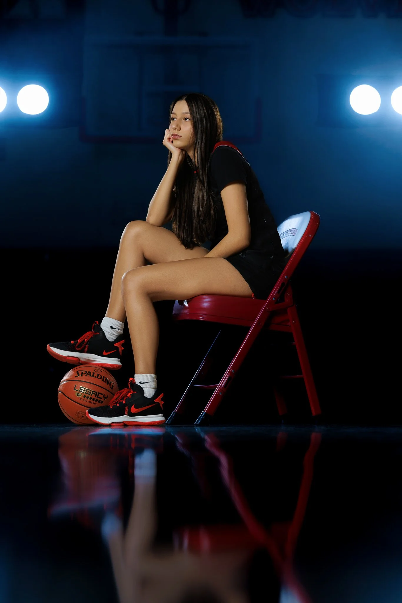 A teenage girl sitting on a red chair in a basketball gym, resting her chin on her hand, with a basketball under her foot, wearing black athletic clothes and Nike sneakers.