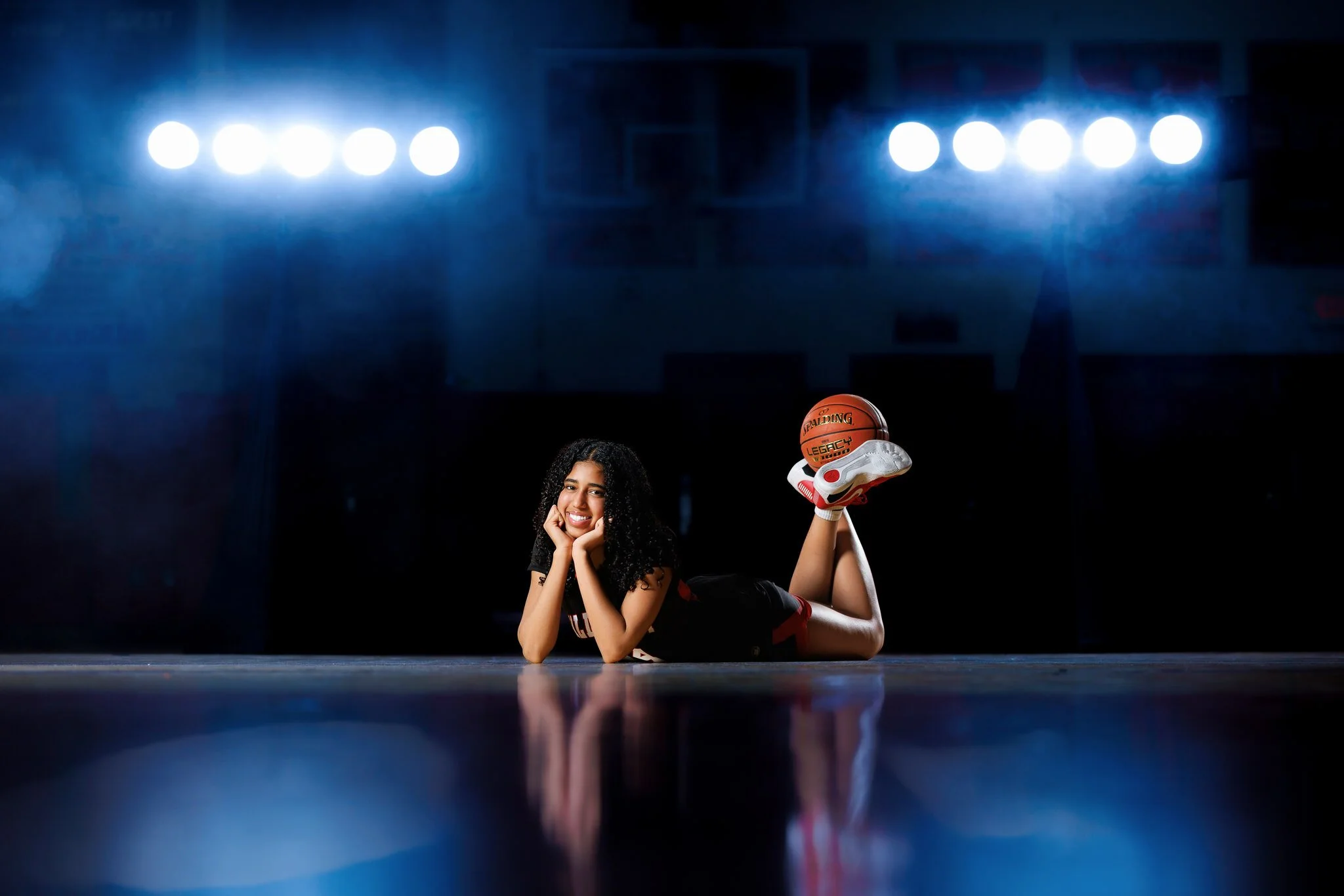 A young female basketball player with curly hair lying on the sports court, smiling, holding a basketball, under bright overhead stadium lights.