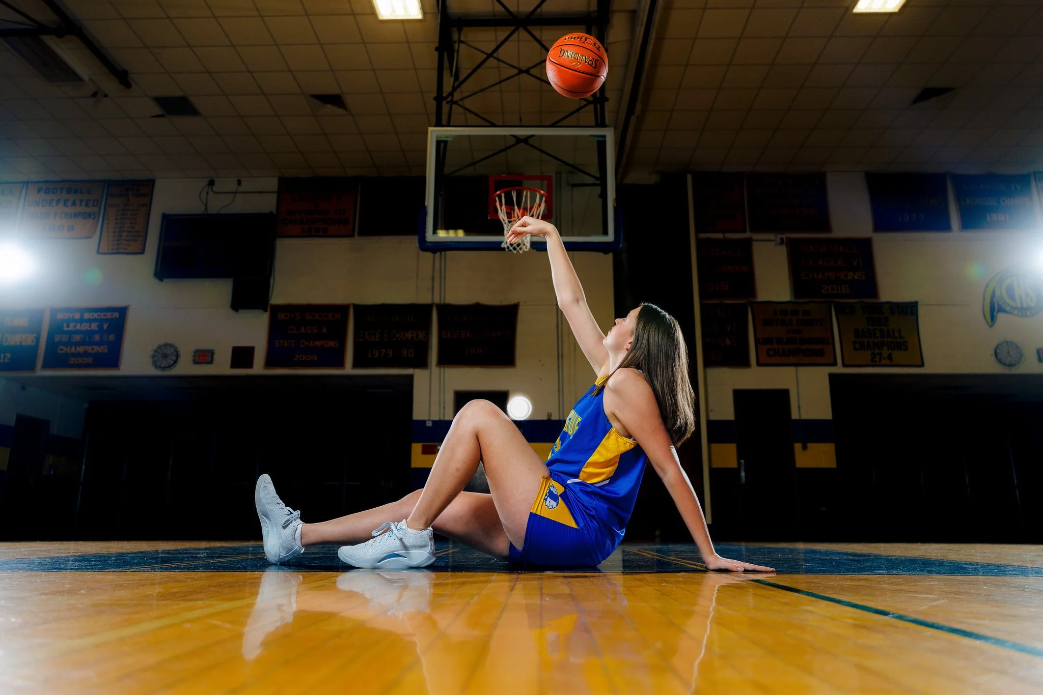 A female basketball player in a blue and yellow uniform sitting on the basketball court floor, shooting a basketball towards the hoop.
