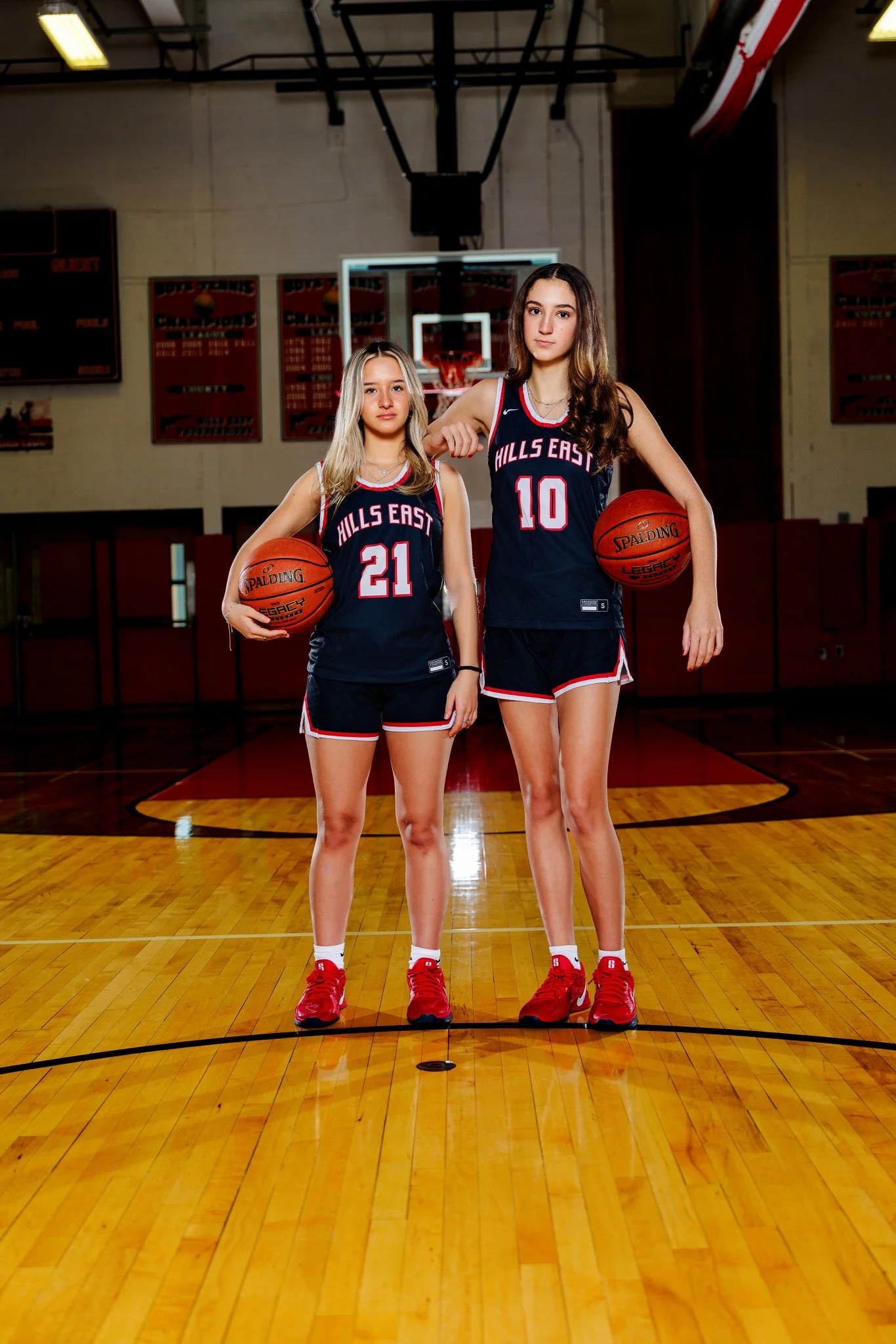 Two female basketball players in navy uniforms with red accents standing on a basketball court, each holding a basketball. The player on the left wears number 21, and the player on the right wears number 10. They are in a gym with a basketball hoop a