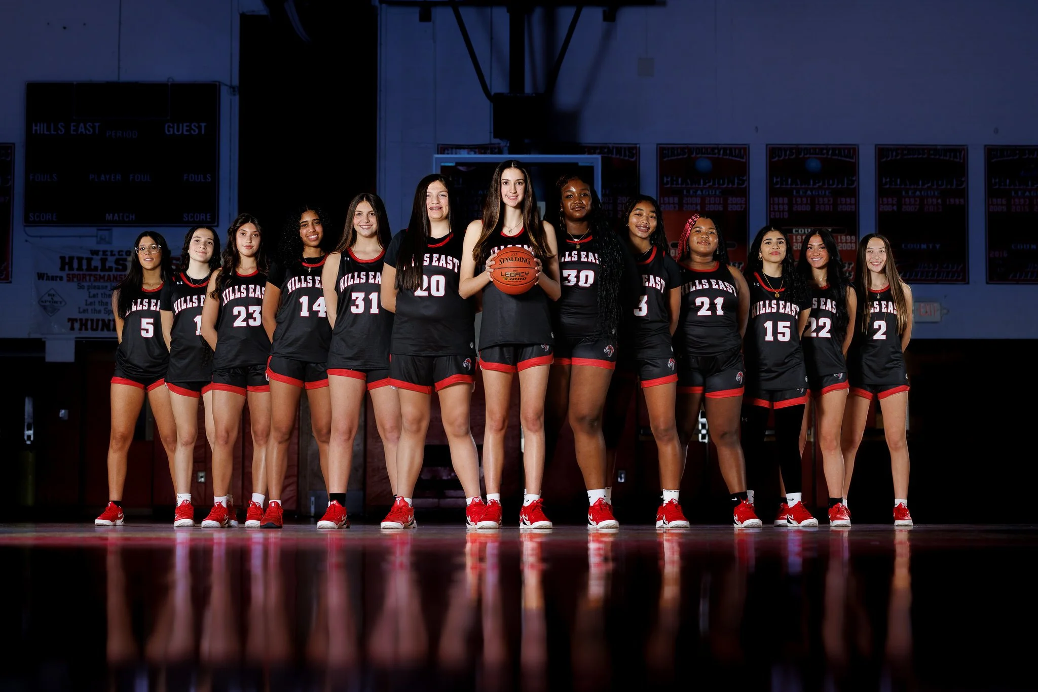A girls' basketball team stands in a line in a gym, wearing black and red uniforms. One girl in the center holds a basketball. The gym background includes scoreboard and banners.