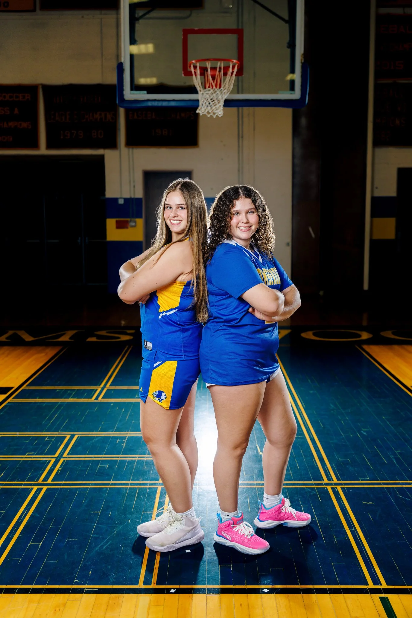 Two young women standing back to back on a basketball court, smiling, with a basketball hoop overhead and banners on the wall in the background. They are dressed in athletic clothing, one in a blue and yellow sports uniform and the other in a blue sh