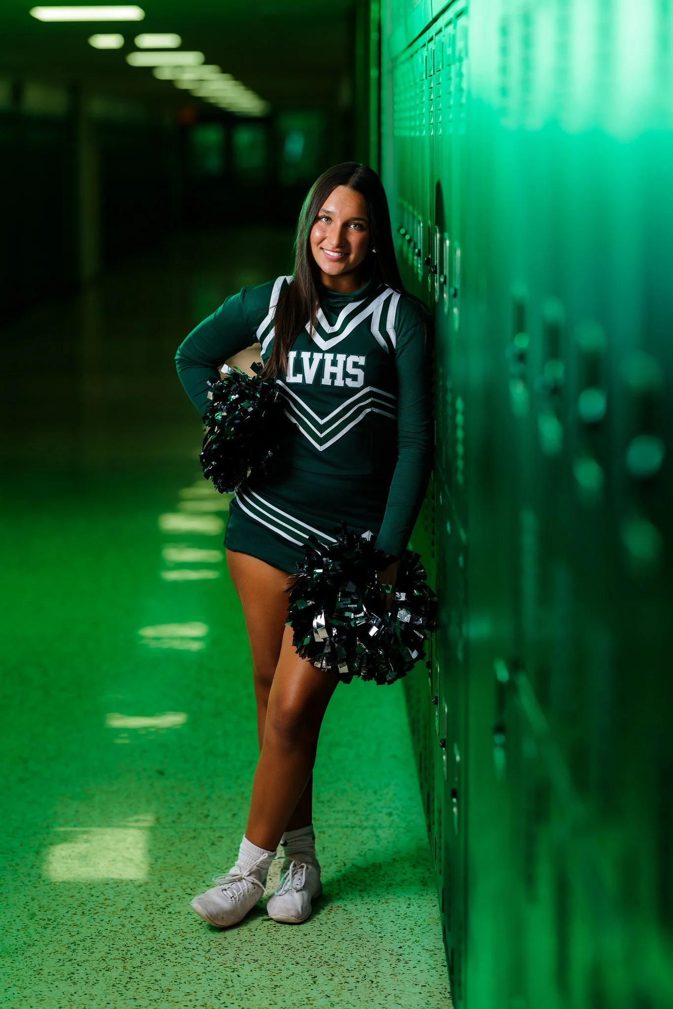 A young female cheerleader in a green and black LVHS cheerleading uniform posing in a school hallway with green lockers.