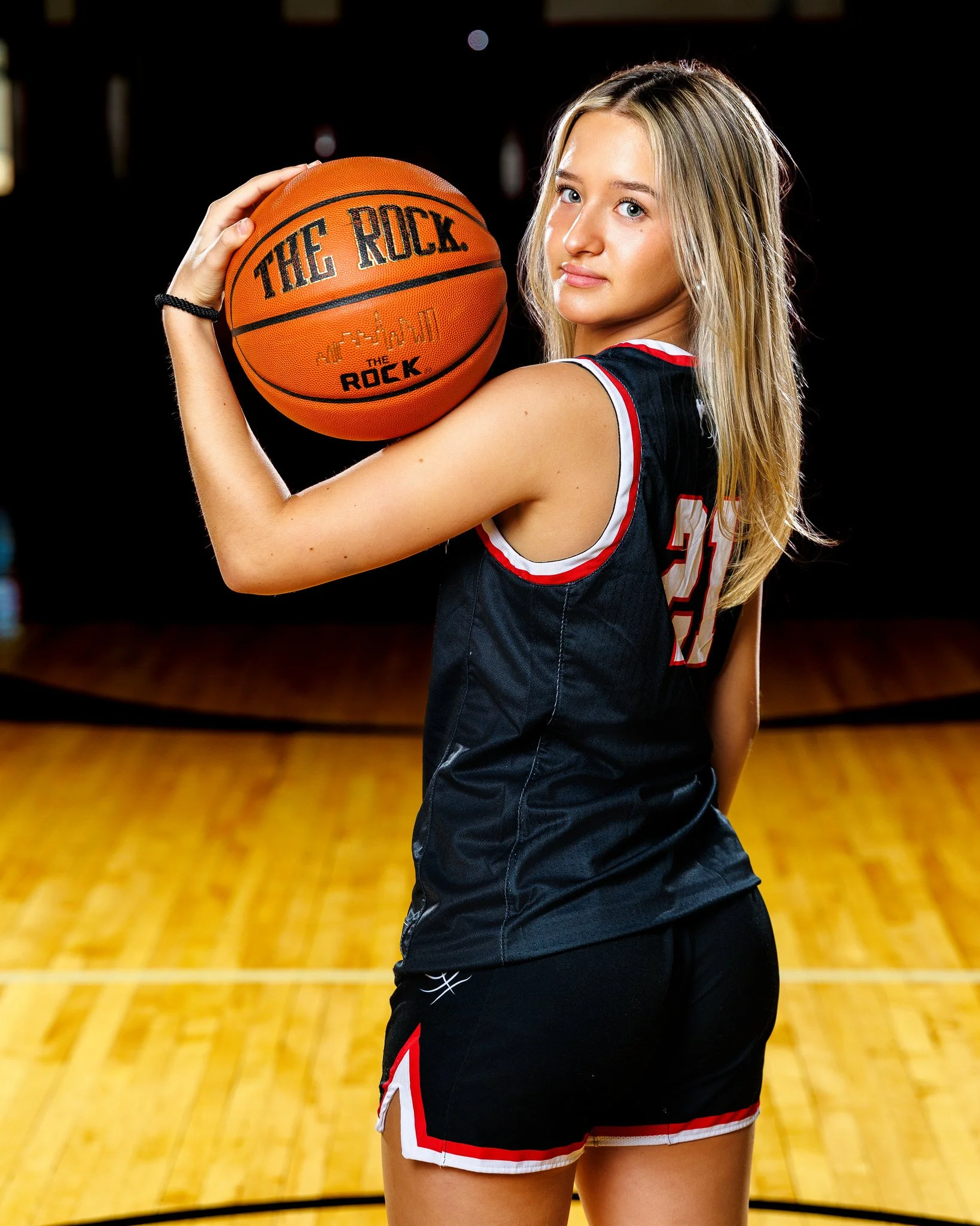 A young woman in a black basketball uniform holding an orange basketball with the words 'THE ROCK' in a gymnasium.
