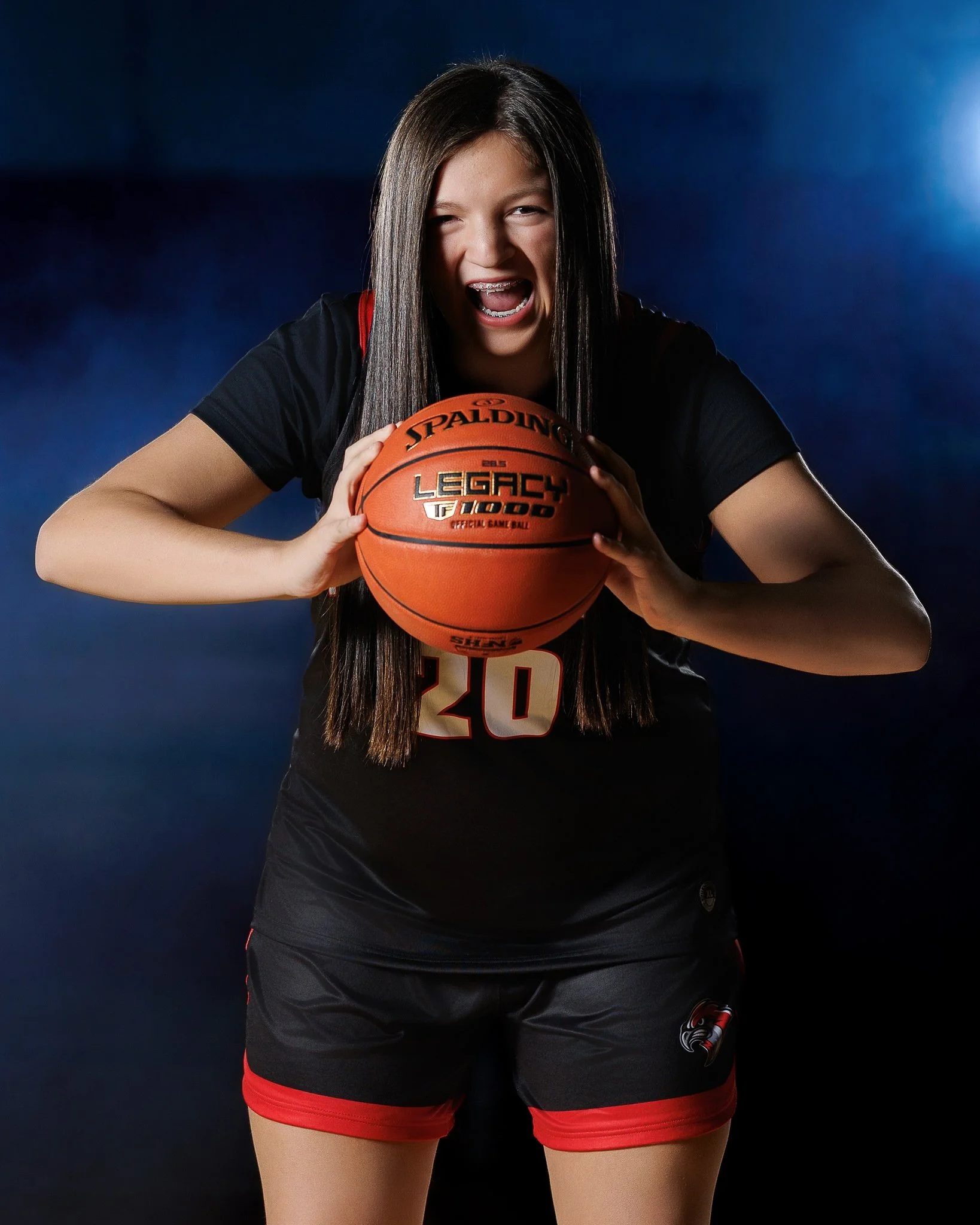 A young female basketball player in a black jersey holding a basketball and posing aggressively on a dark background.