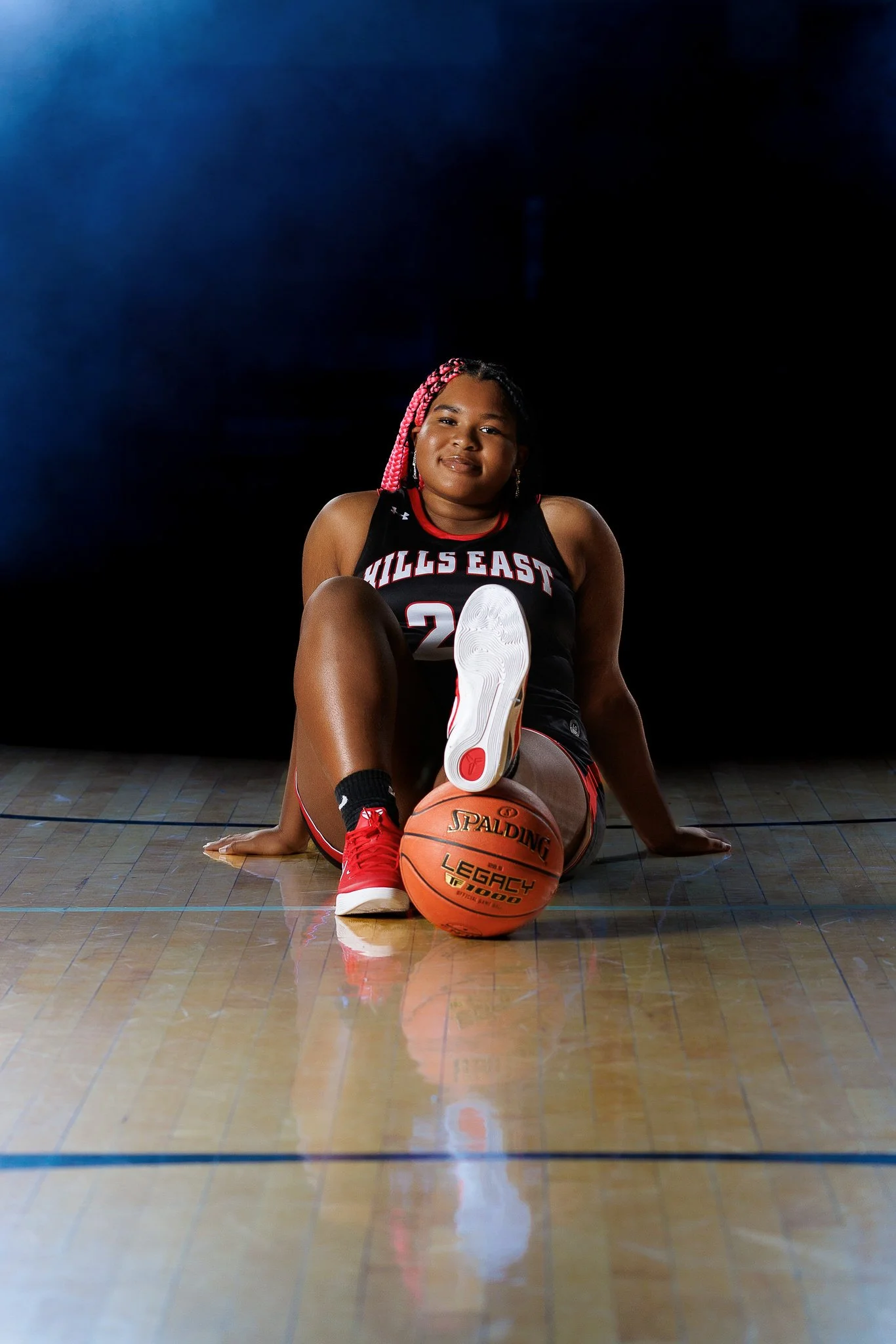A young female basketball player sitting on a basketball court, with one foot on a basketball and the other raised, wearing a black jersey with red and white accents, and pink braids in her hair.