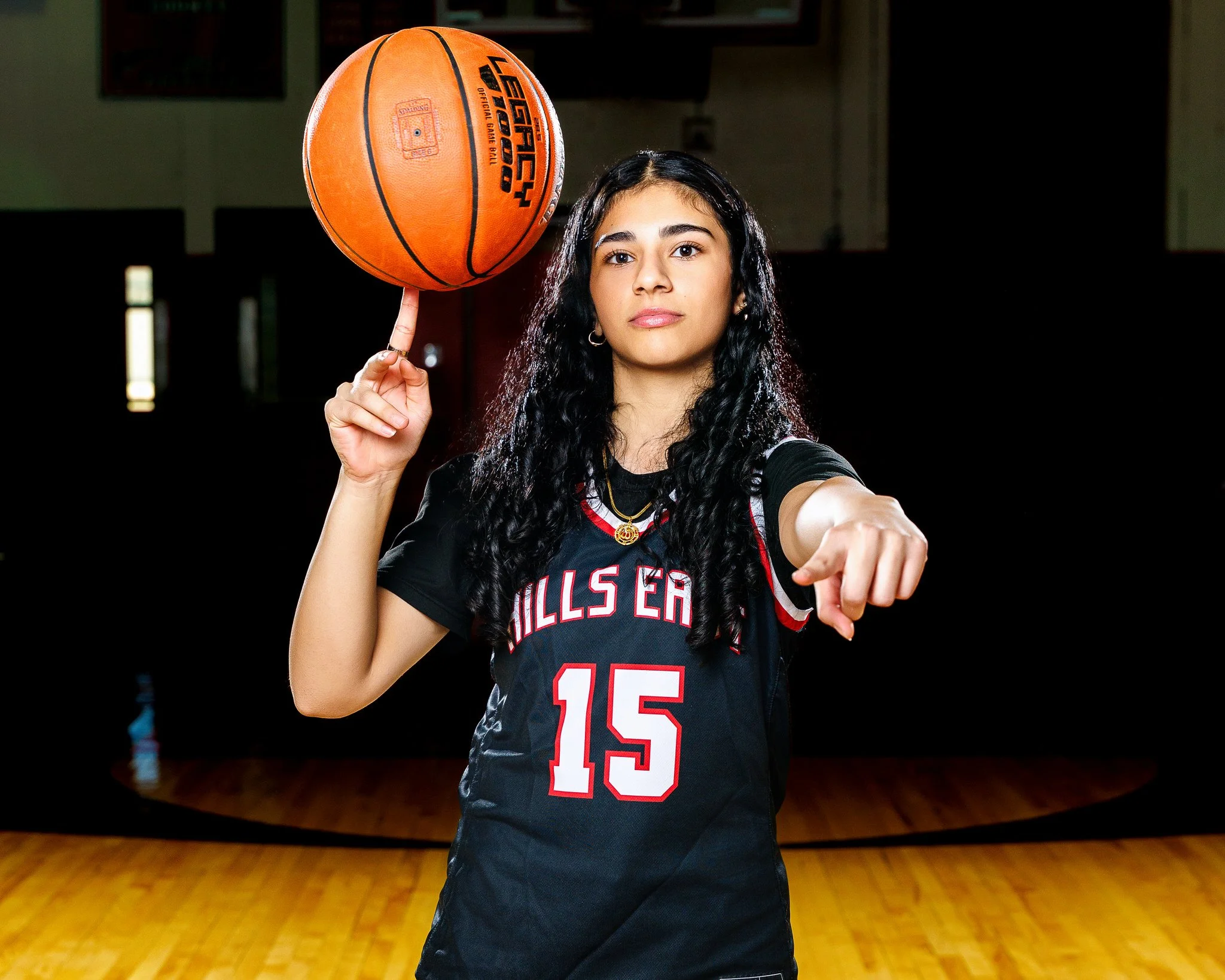 A young woman with long curly black hair posing in a basketball gym wearing a black sports jersey with the number 15, spinning a basketball on her finger, and pointing forward.