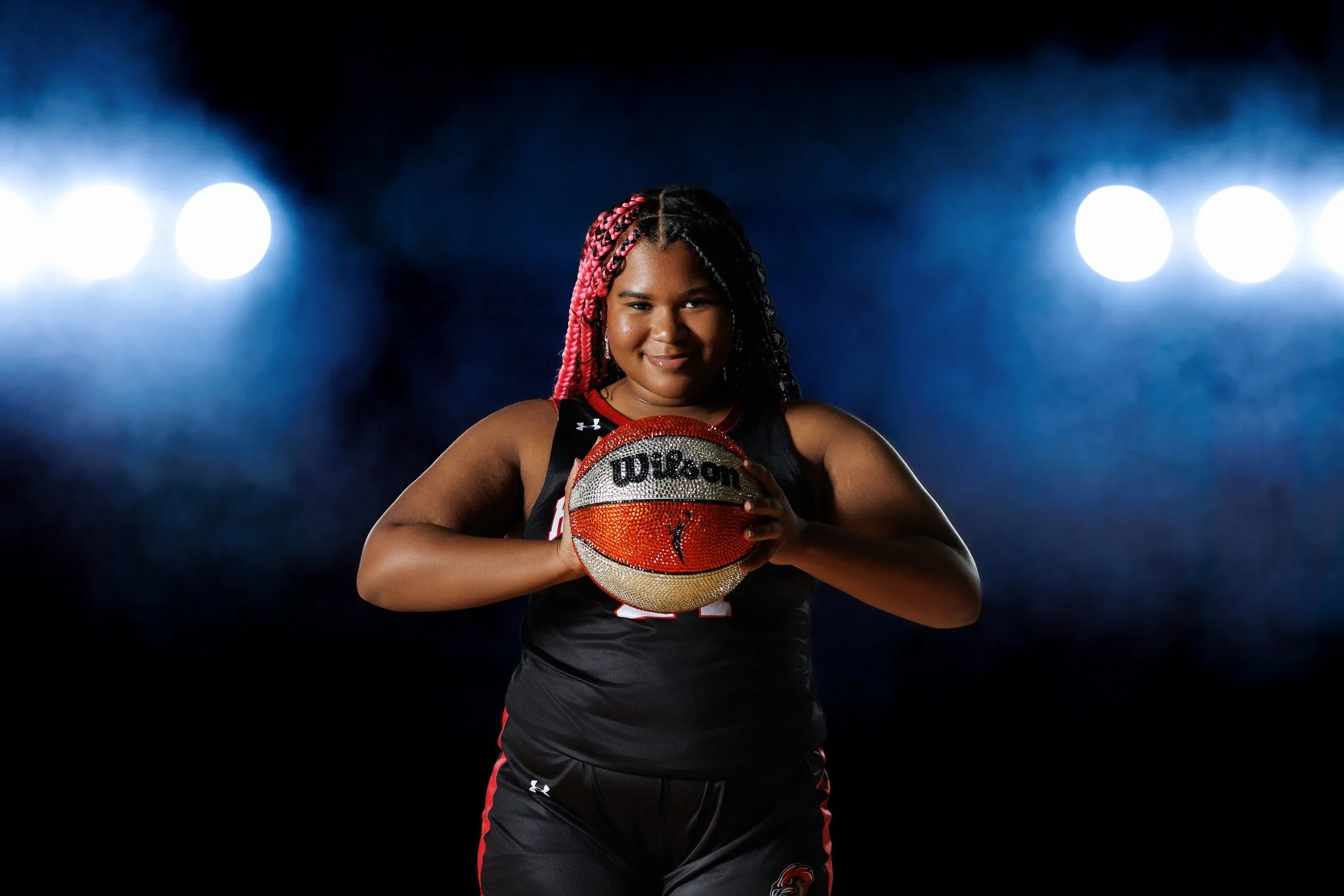 A female basketball player with red and black braids, holding a diamond-studded Wilson basketball, standing in a dark studio with bright lights behind her.