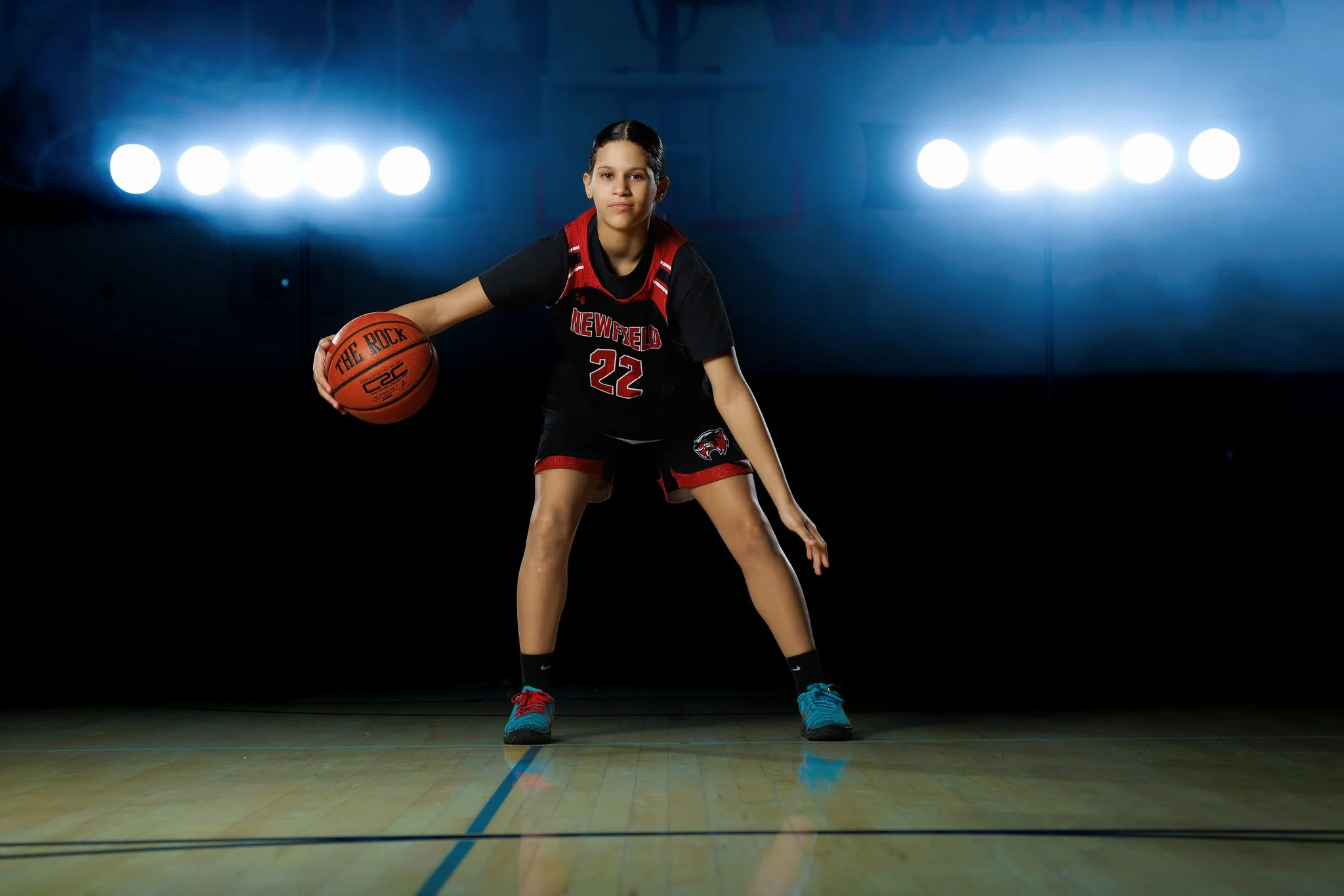 Young female basketball player in black and red uniform holding basketball in a gym, with bright lights in the background.