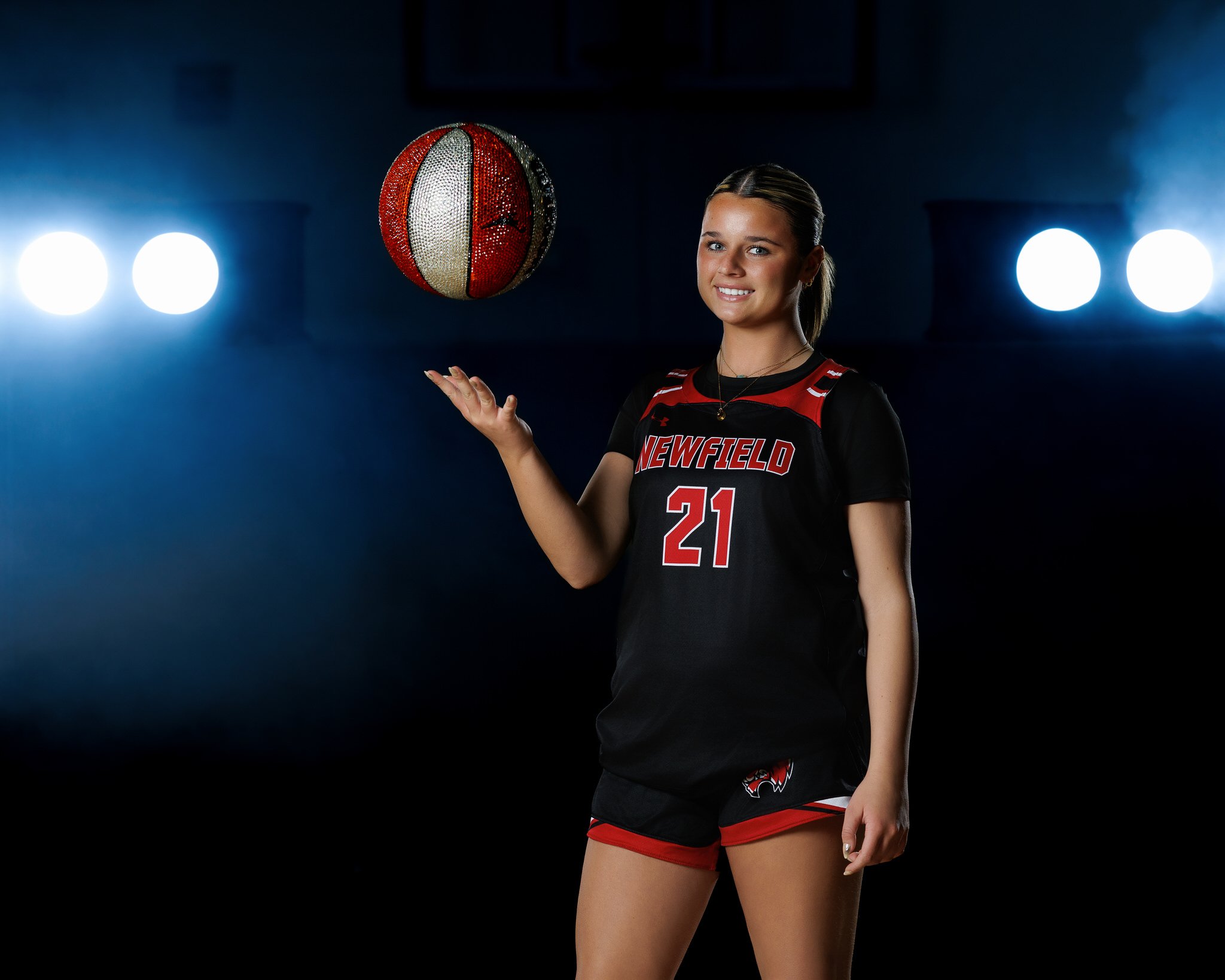 A young female volleyball player wearing a black and red uniform with the number 21, holding a sparkly volleyball, standing in a sports arena with bright lights.