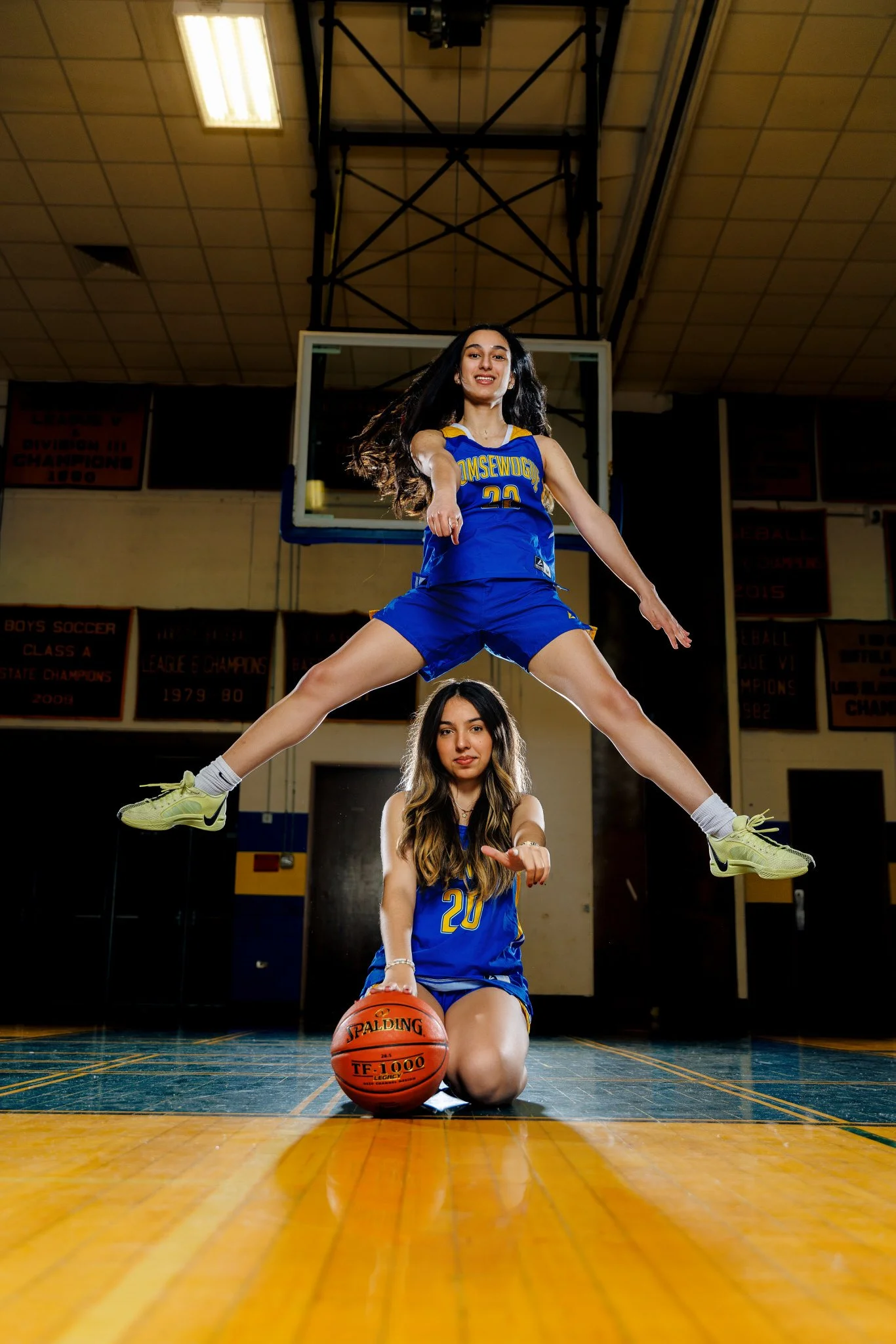 Two young female basketball players in blue uniforms inside a gymnasium. One player is sitting on the floor holding a basketball, while the other is leaping in the air above her, with legs spread apart and arms extended.