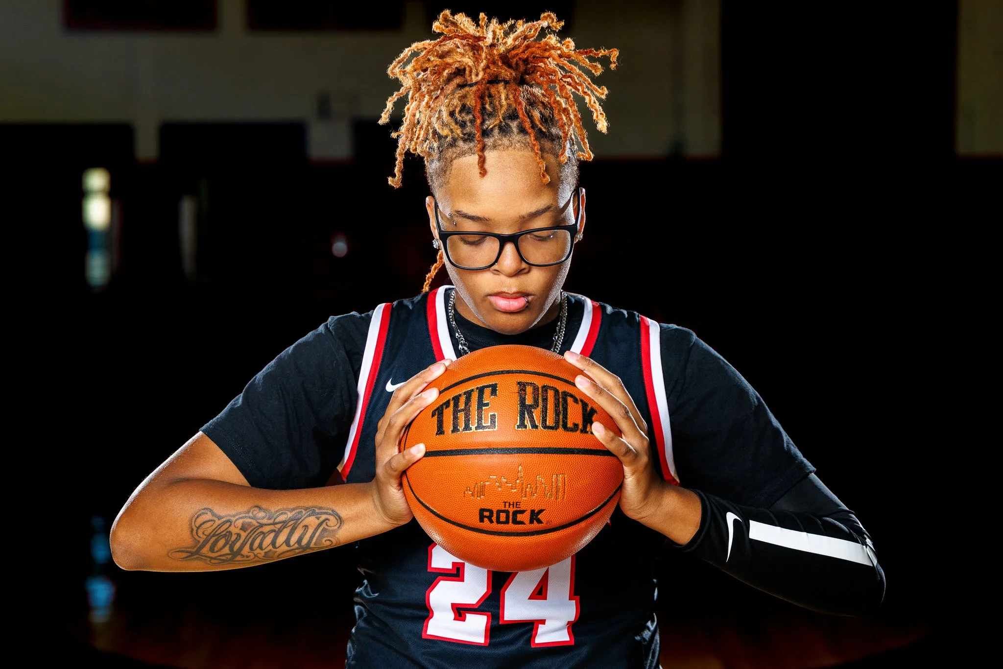 A female basketball player with pink dreadlocks, glasses, and tattoos on her arm, wearing a black jersey with red and white accents, holding a basketball with both hands, standing in a dimly lit basketball court.