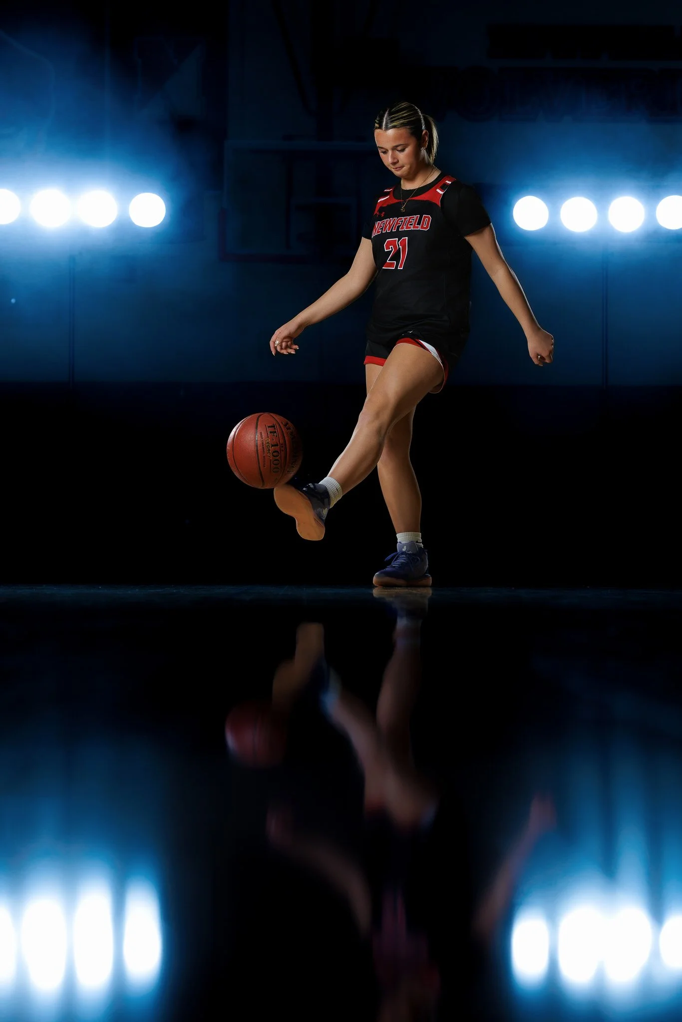 Young female basketball player in a black uniform kicks a basketball on a dark indoor court with bright spotlights behind her.