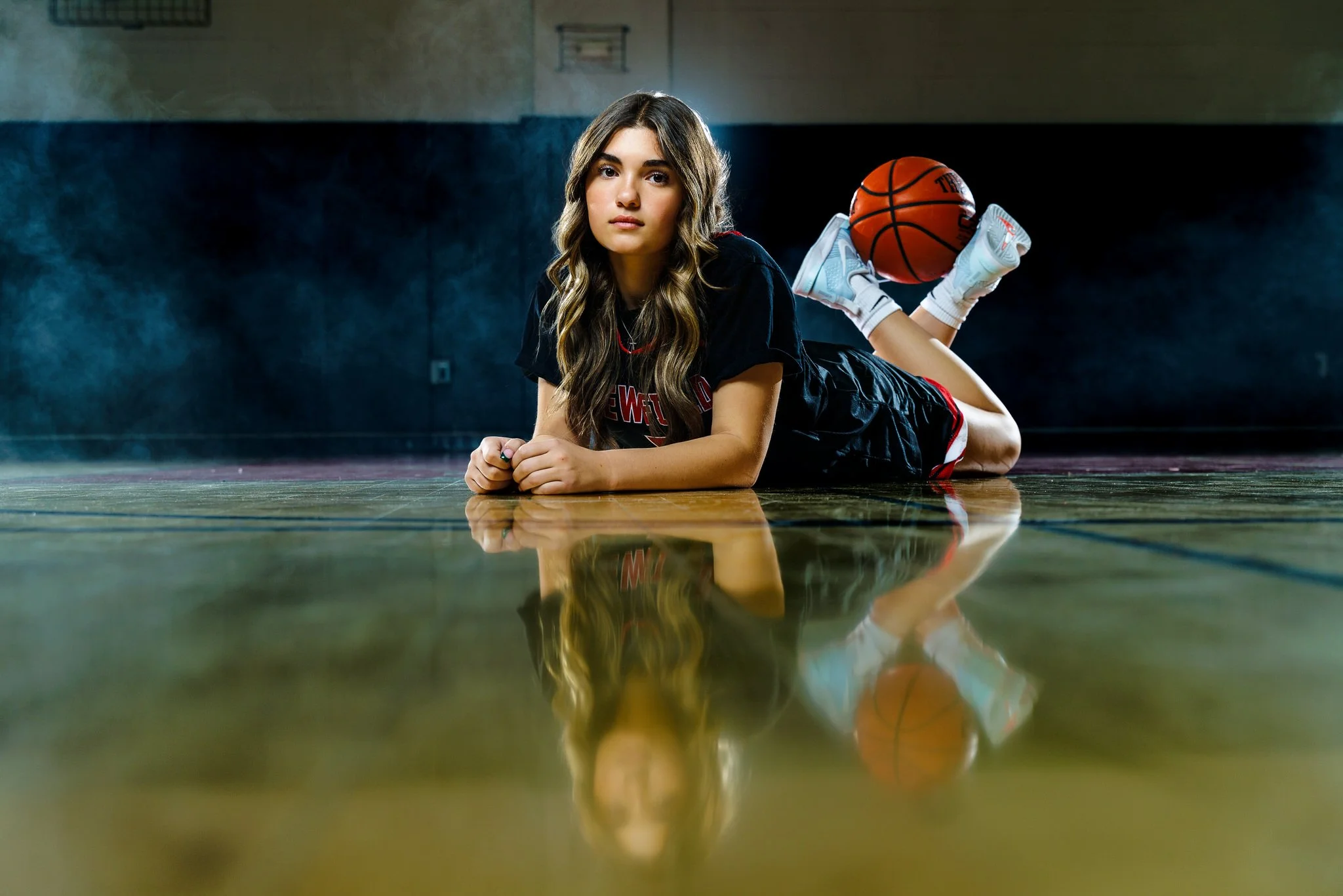 A young woman with long wavy hair lying on the gym floor, wearing a black sports jersey, with her hands clasped and her chin resting on them. She is in a gymnasium with a basketball behind her, propped up on her feet, which are in white sneakers. The