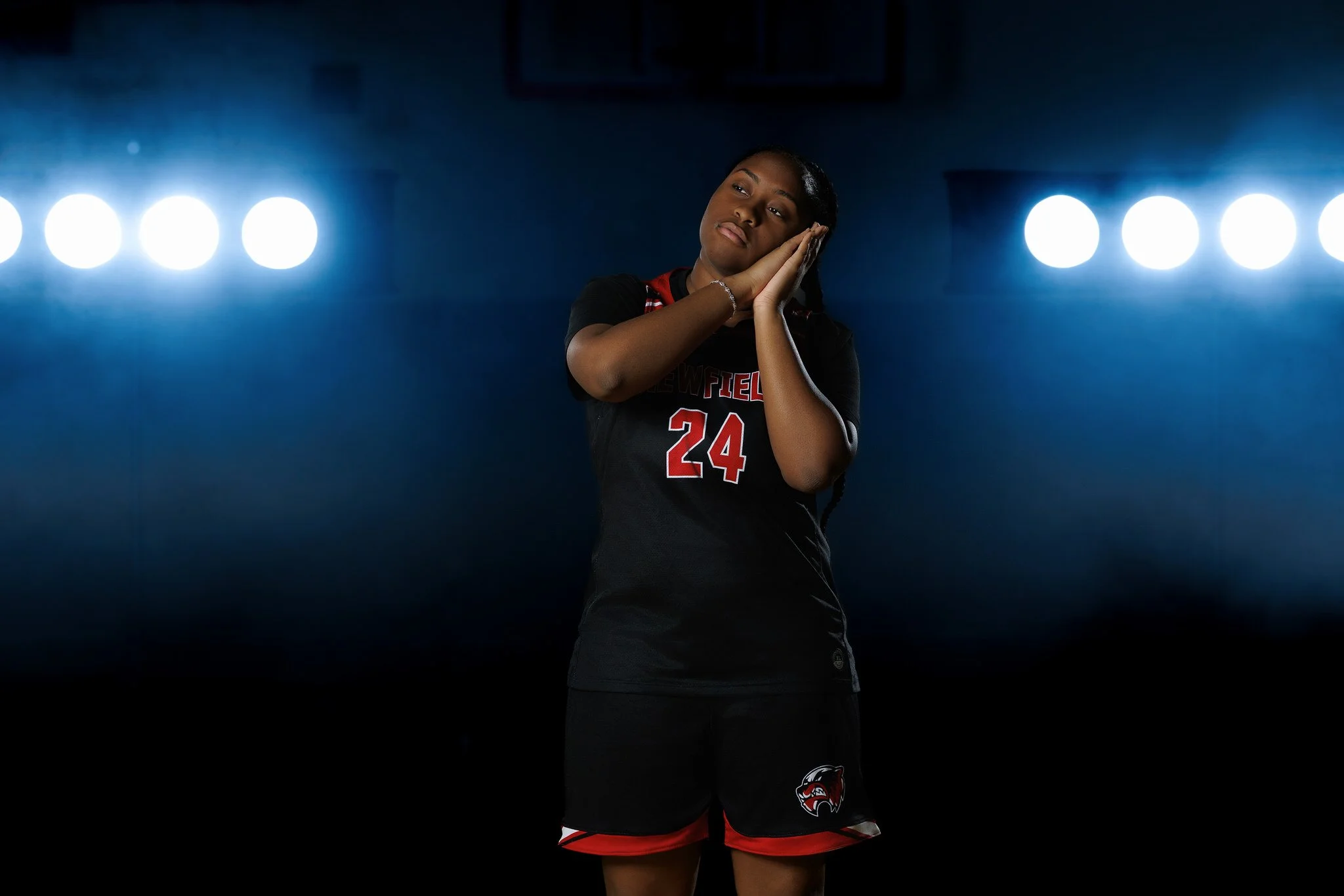 A female athlete in a black sports jersey with the number 24, standing with her hands on her cheeks, looking contemplative against a dark background with bright overhead lights.