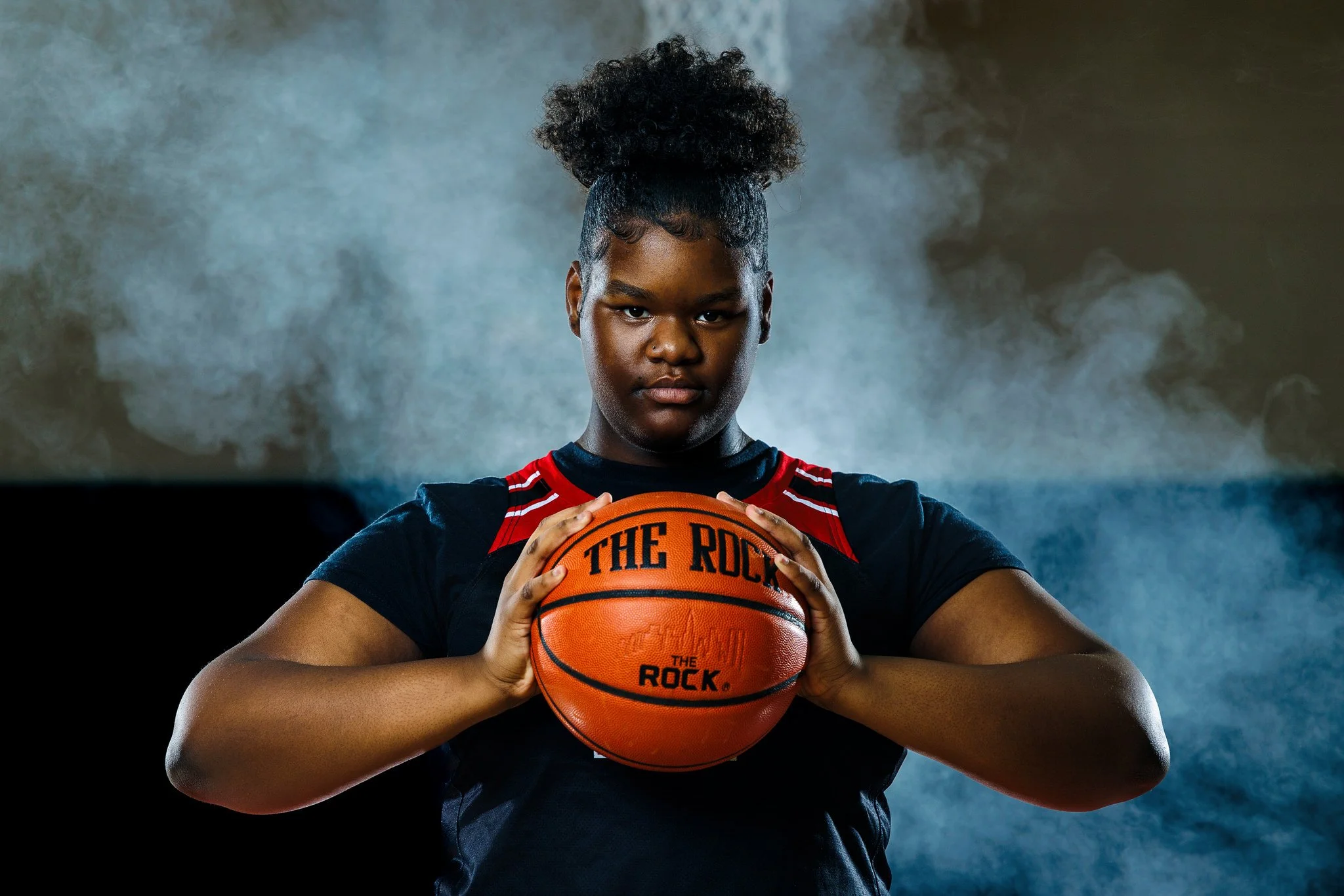 A young female basketball player in a black and red uniform holding a basketball with both hands, looking serious, with dark smoke or mist in the background.