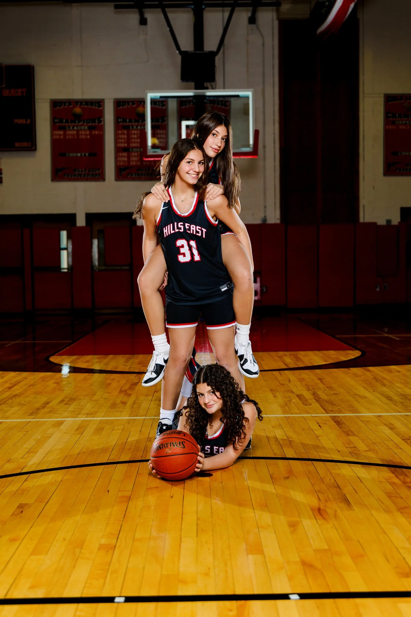 Three young girls in basketball uniforms on a basketball court, with two girls standing and one girl lying on the floor holding a basketball.