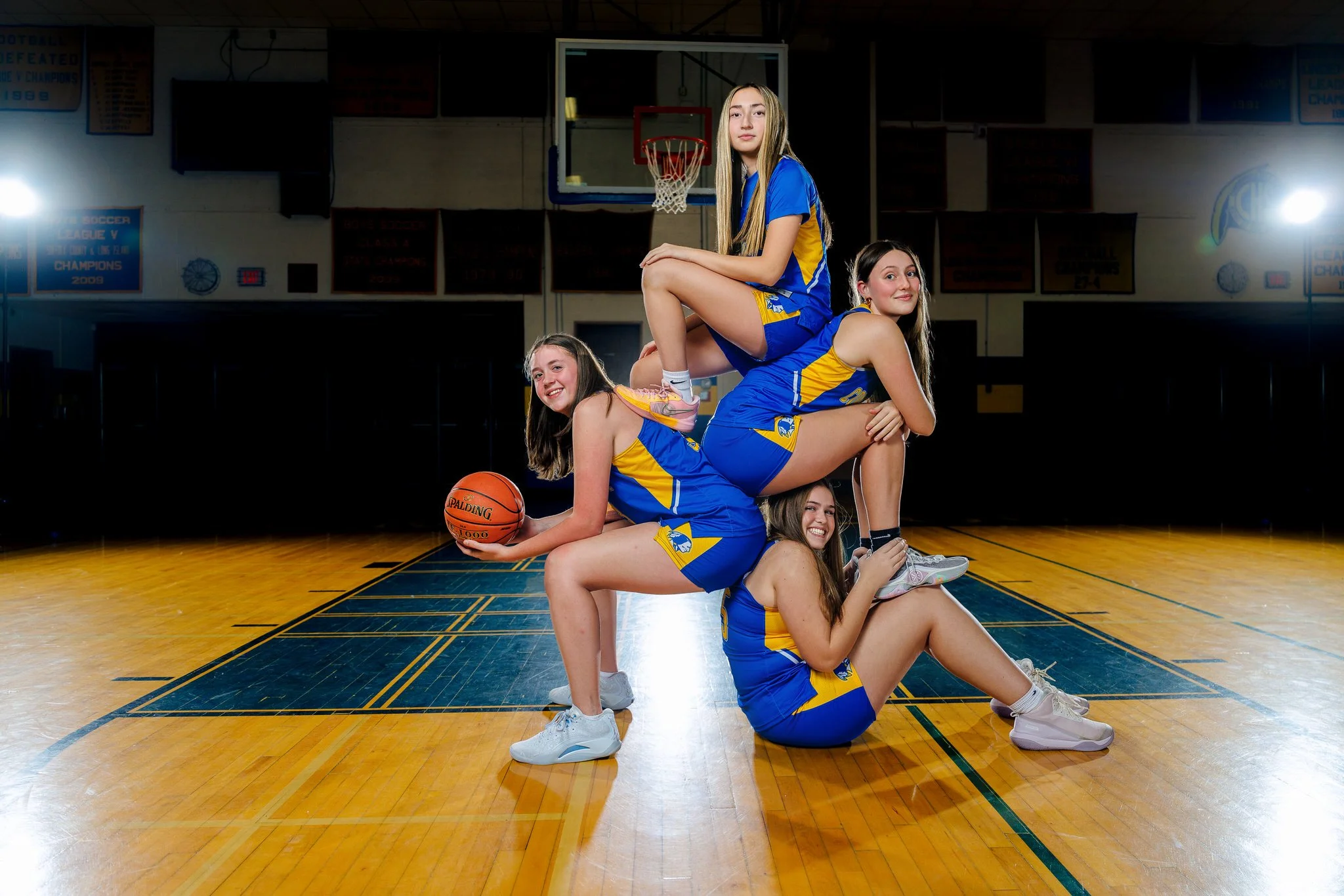 Four young female basketball players in blue and yellow uniforms stacked in a pyramid pose on an indoor basketball court with one holding a basketball.