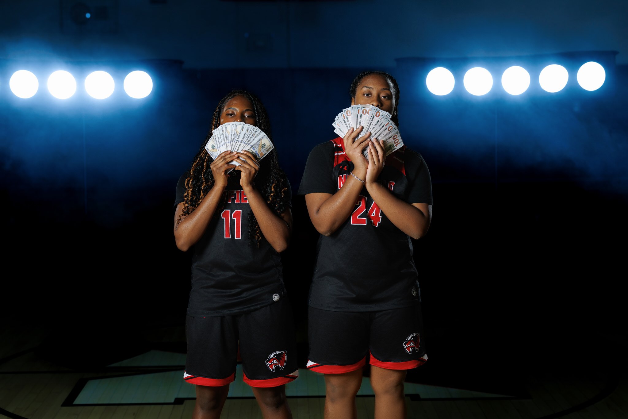 Two female basketball players in black jerseys with red trim, holding large fans of money, standing on a basketball court with bright overhead lights.