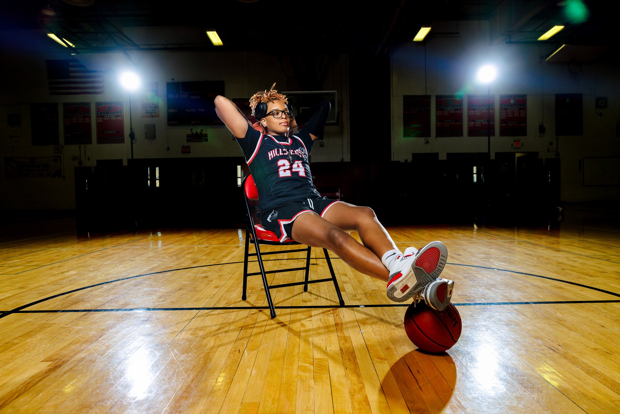 A female basketball player with dreadlocks, glasses, and a black jersey with the number 24, sitting on a red chair in a gymnasium with her arms behind her head, a basketball resting on the court beneath her, and spotlights overhead.