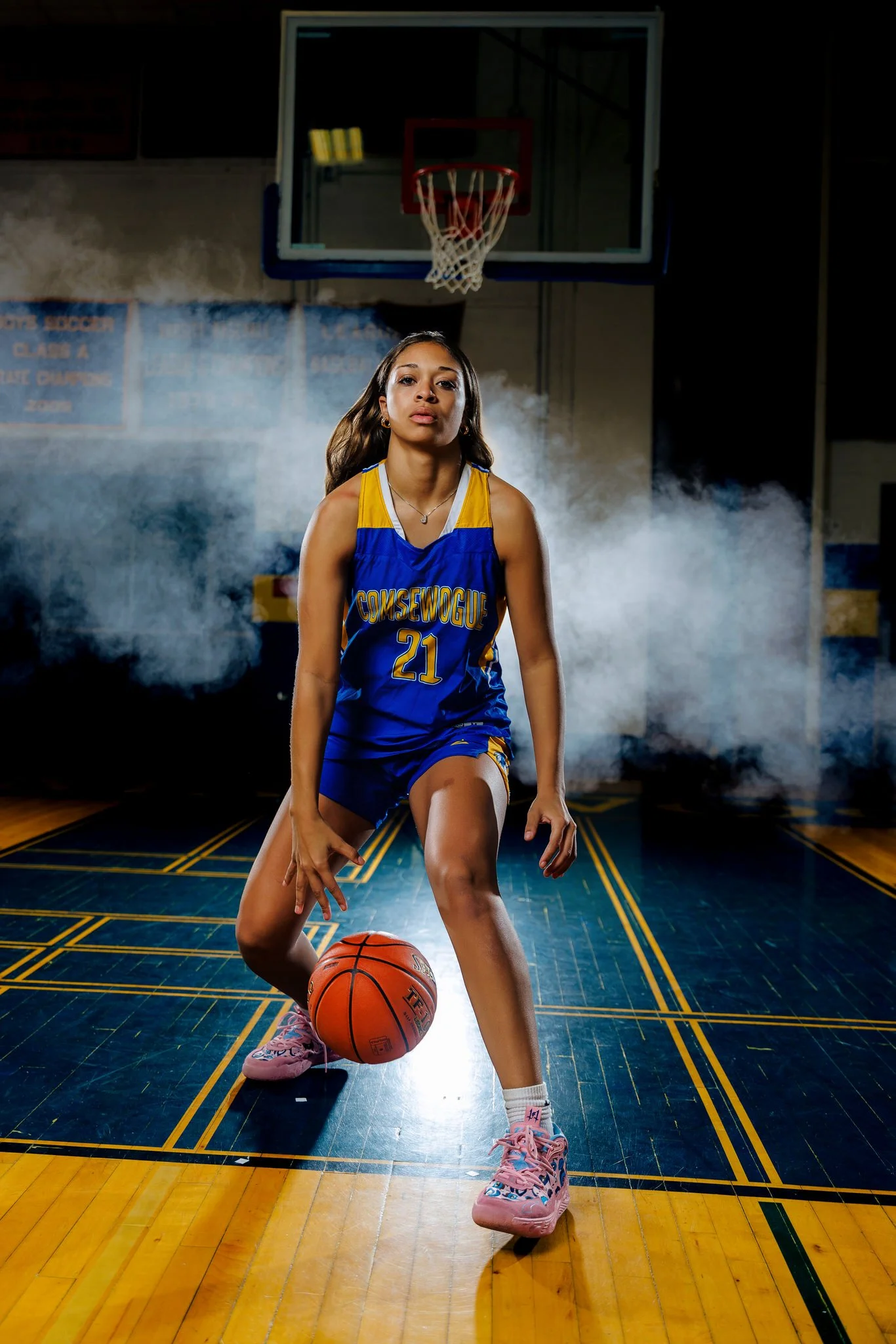 A young female basketball player in a blue and yellow jersey with the number 21, dribbling a basketball on an indoor court with a basketball hoop and backboard overhead, and a smoky background.
