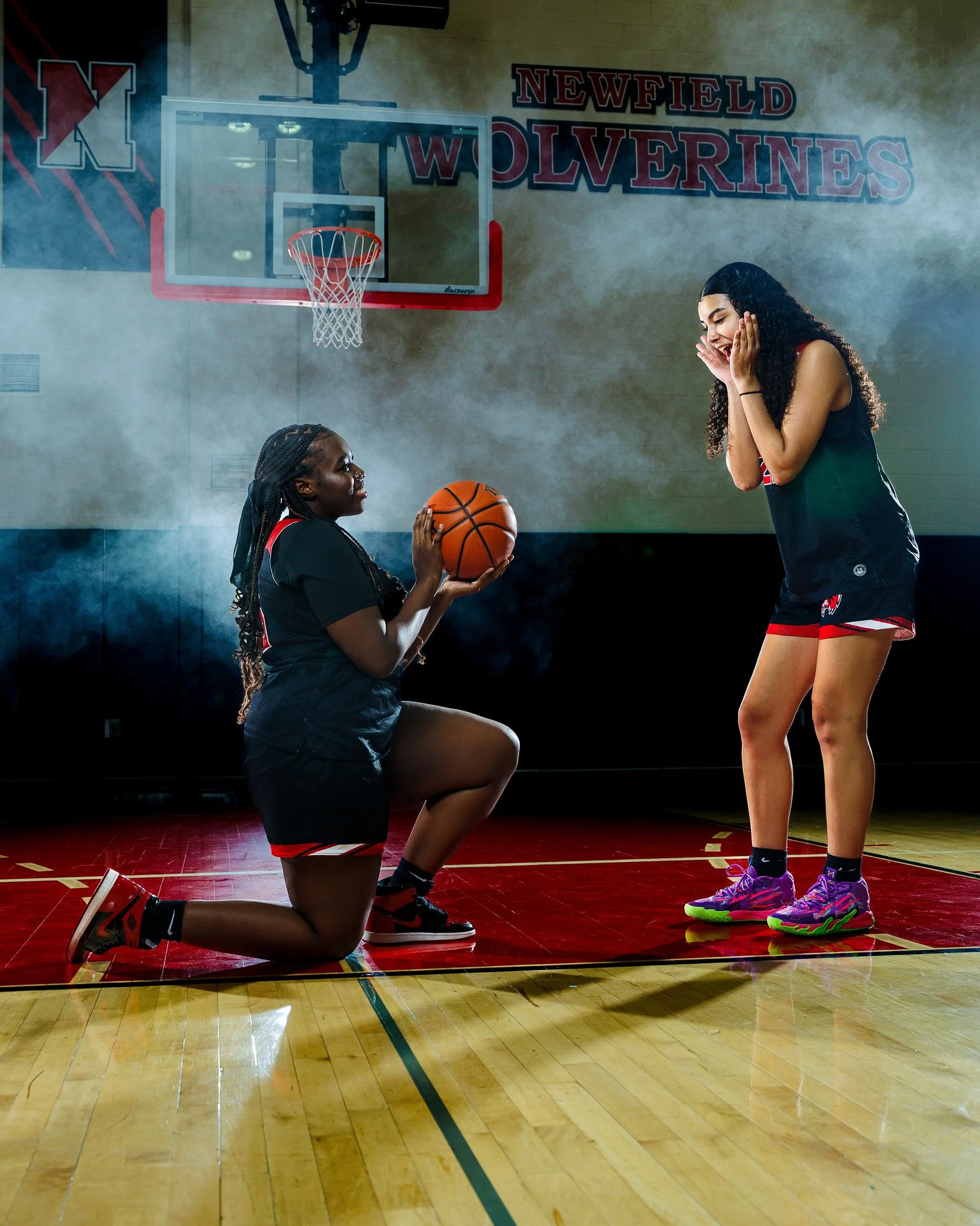 Two women in basketball uniforms on an indoor court, one kneeling and holding a basketball, the other standing with hands on her face, in front of a basketball hoop and sign that reads "Newfield Wolverines".