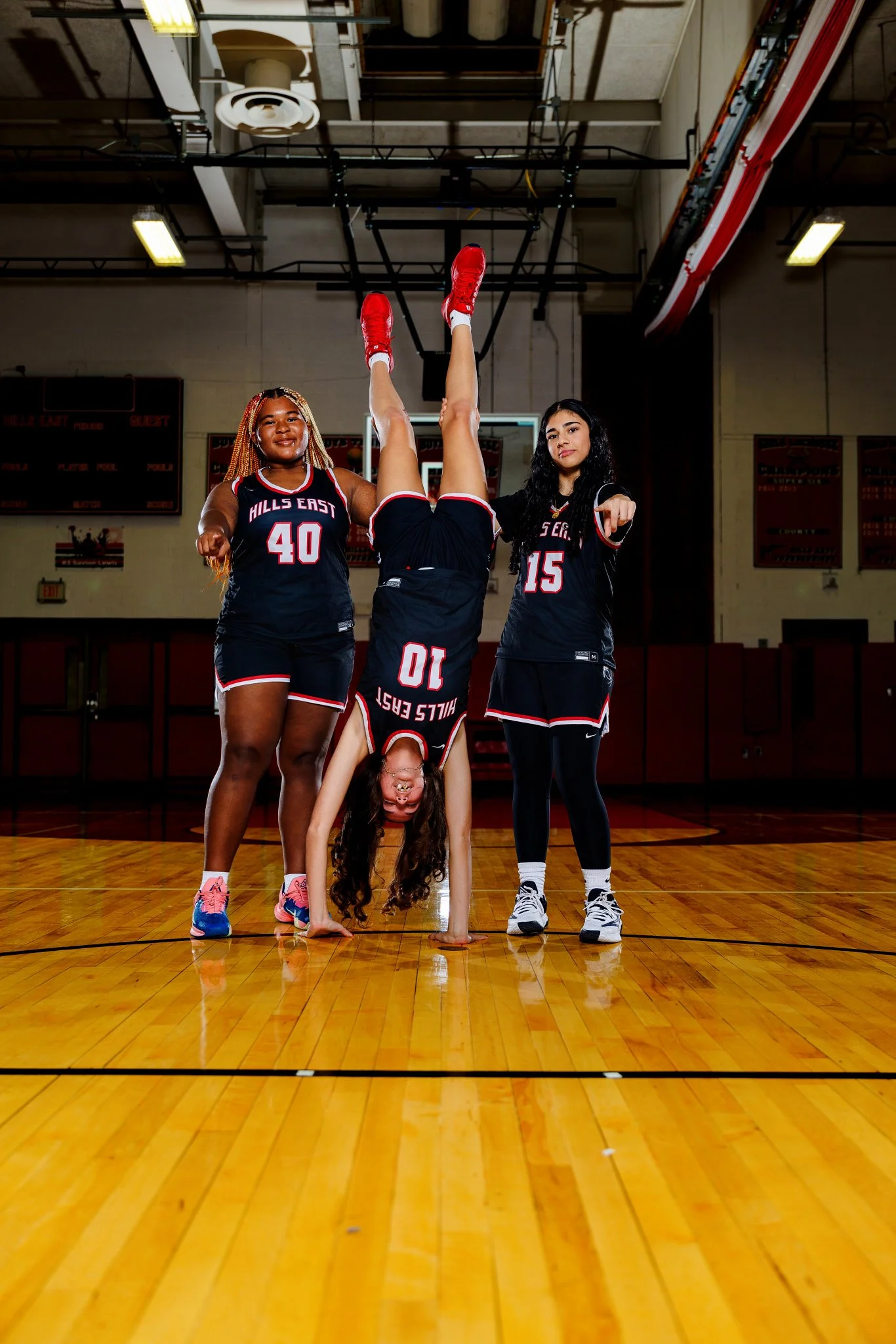 Three young women in basketball uniforms perform a handstand on a basketball court, with one woman upside down with her legs straight up, and the other two standing on either side, pointing towards the camera.