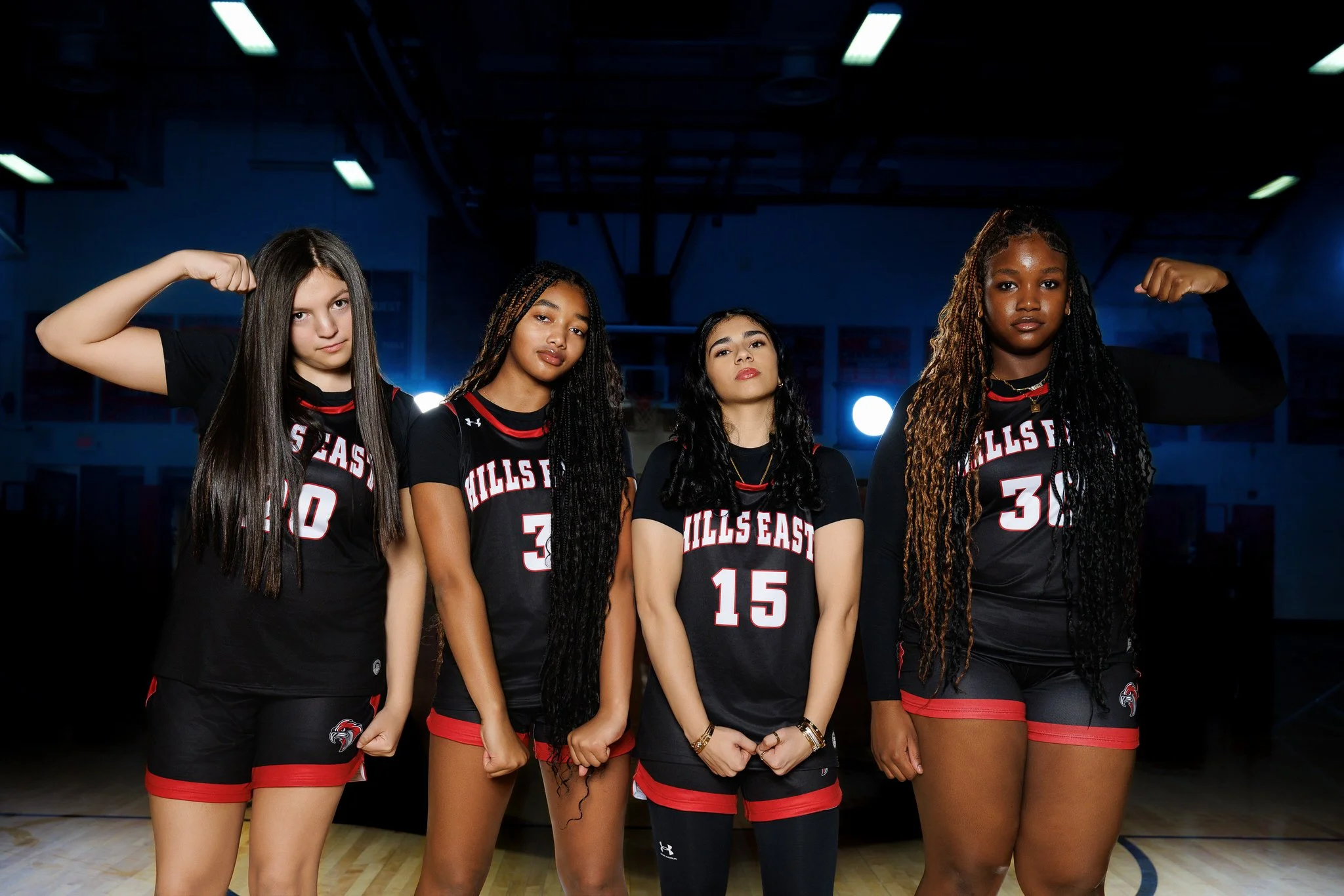 Four young women in basketball uniforms standing on a basketball court inside a gym, striking confident poses.