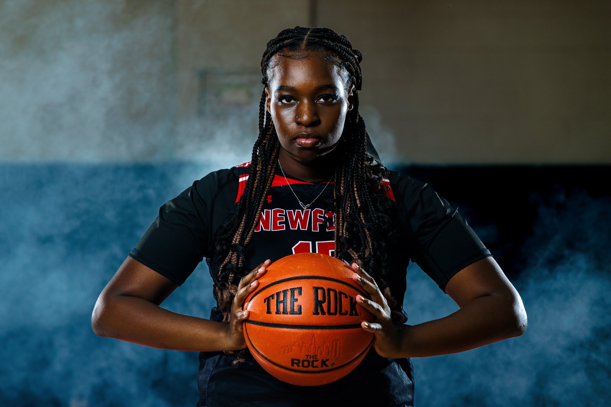A young female basketball player with long braided hair wearing a black and red uniform holding an orange basketball with black lettering in a gym with blue and gray walls.
