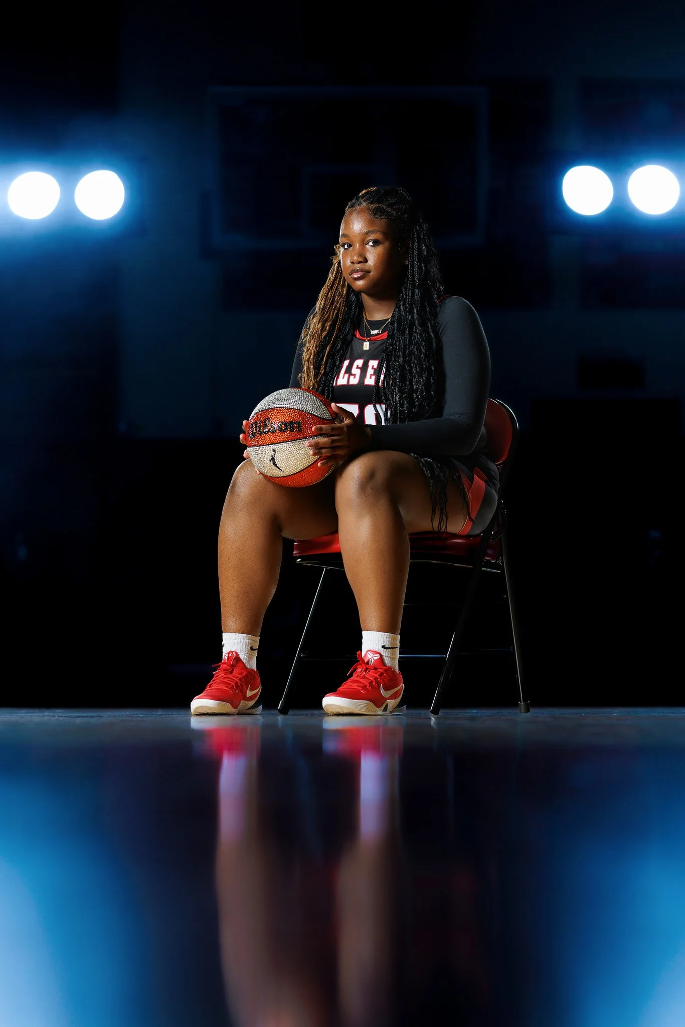 A young woman in a black and red basketball uniform sitting on a chair in a gymnasium, holding a basketball with a lot of rhinestones, under bright overhead lights.