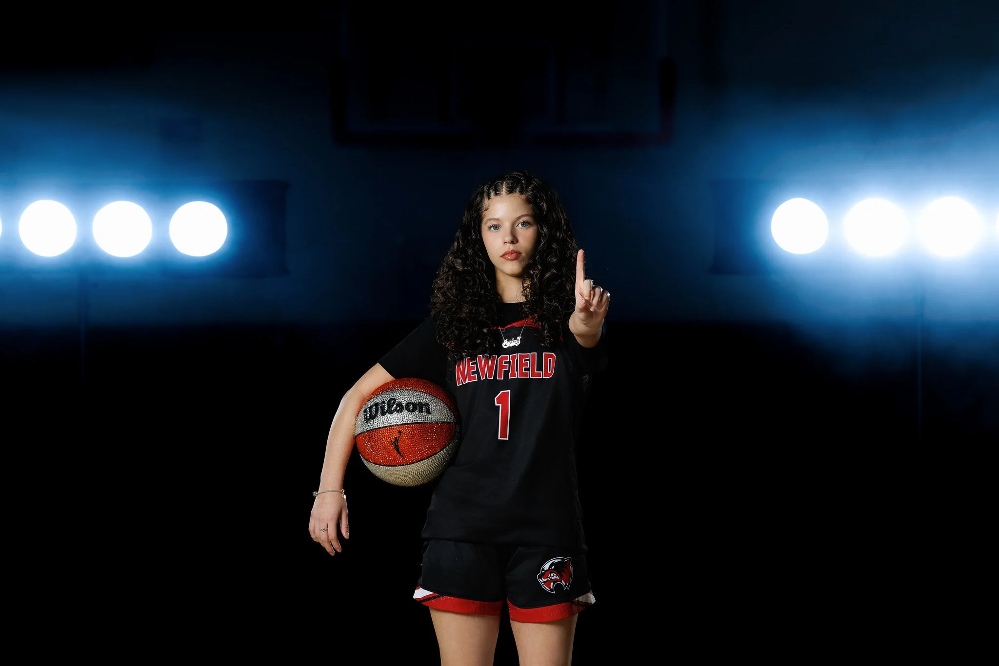 Young female basketball player in black uniform holding a basketball, pointing upward, standing on a dark court with bright lights in the background.