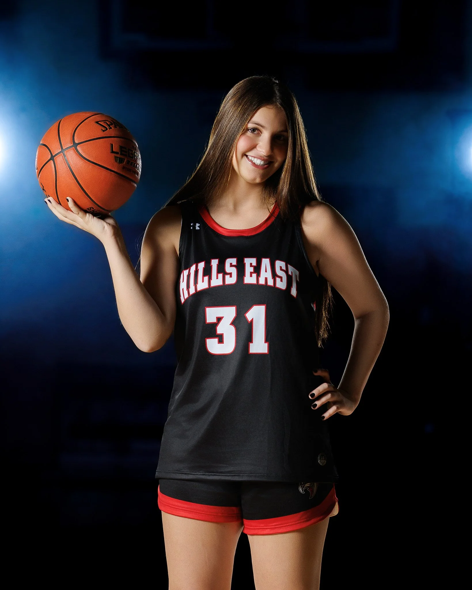 Young woman in a black and red basketball jersey holding a basketball with a smile.