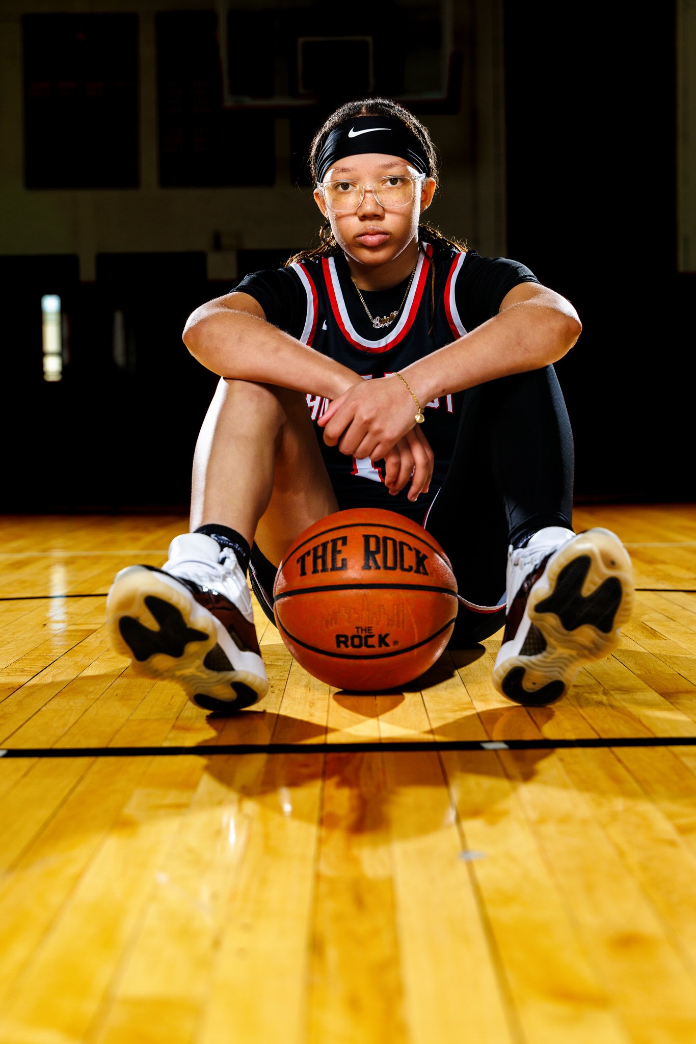 A young female basketball player sitting on a basketball court with a basketball in front of her. She is wearing a black sports uniform, sneakers, glasses, and a headband, and looking directly at the camera.
