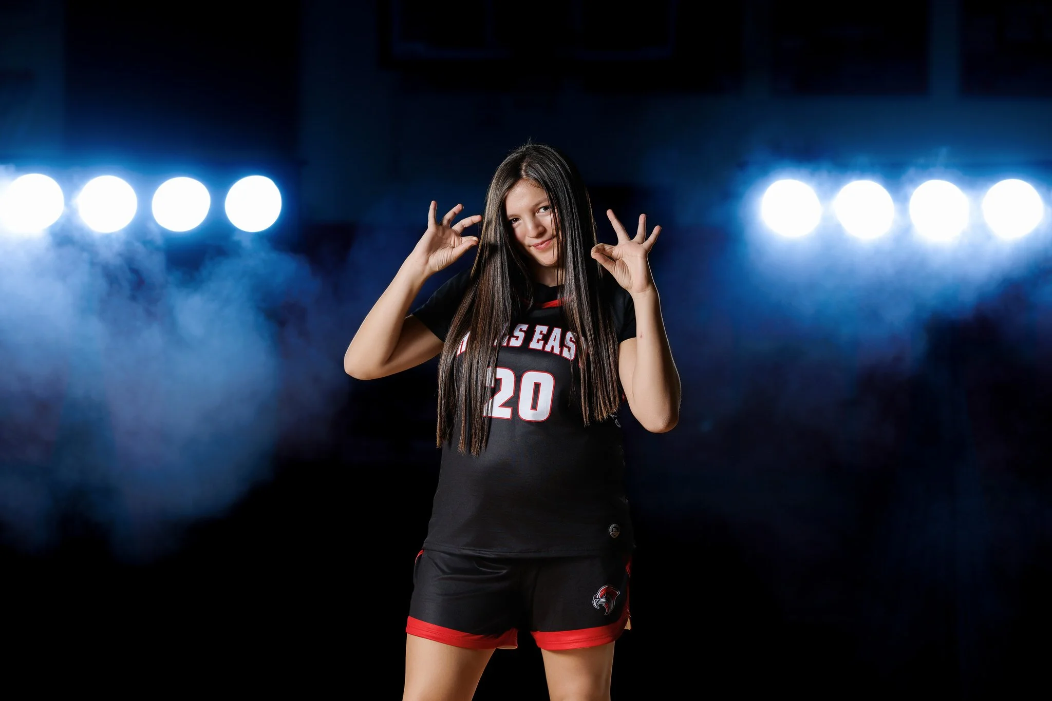Young female volleyball player posing confidently in a black and red uniform with the number 20, against a dark background with bright stage lights and fog.