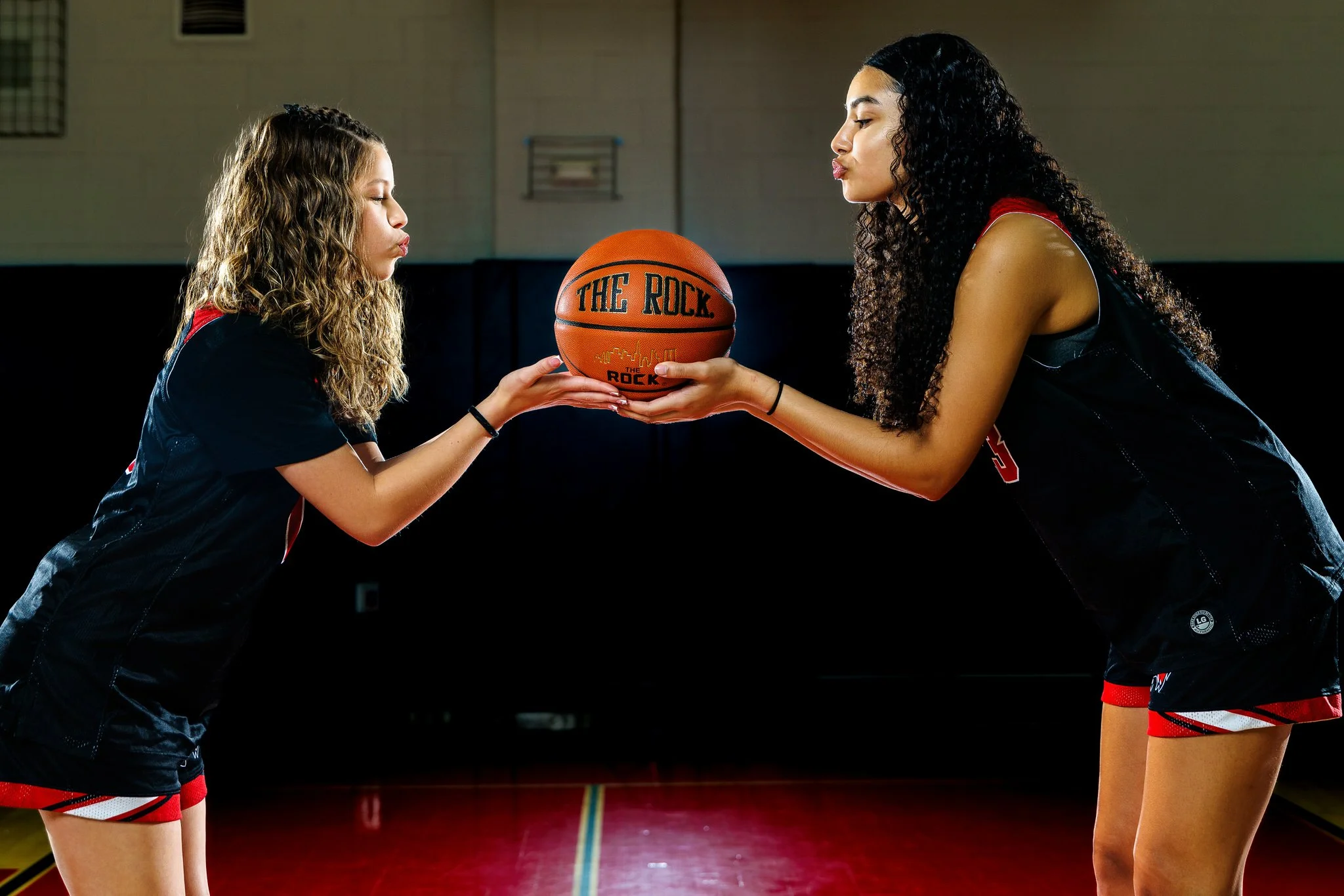 Two female basketball players, one younger and one older, exchanging a basketball on an indoor court.