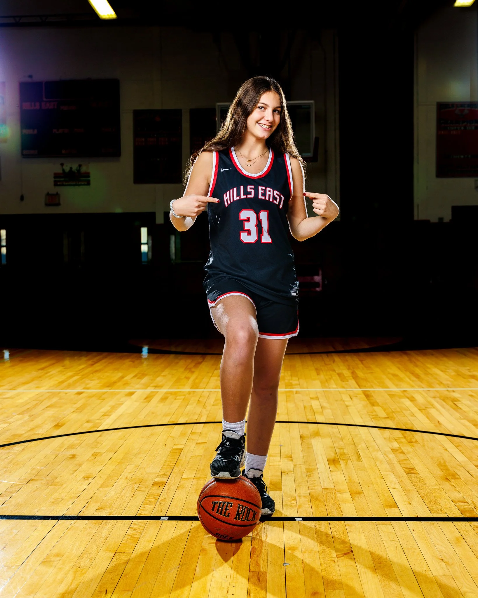 A young woman in a black basketball uniform with red and white trim, numbered 31, pointed at her jersey while standing on a basketball court with one foot on a basketball that has the words "THE ROCK" printed on it.